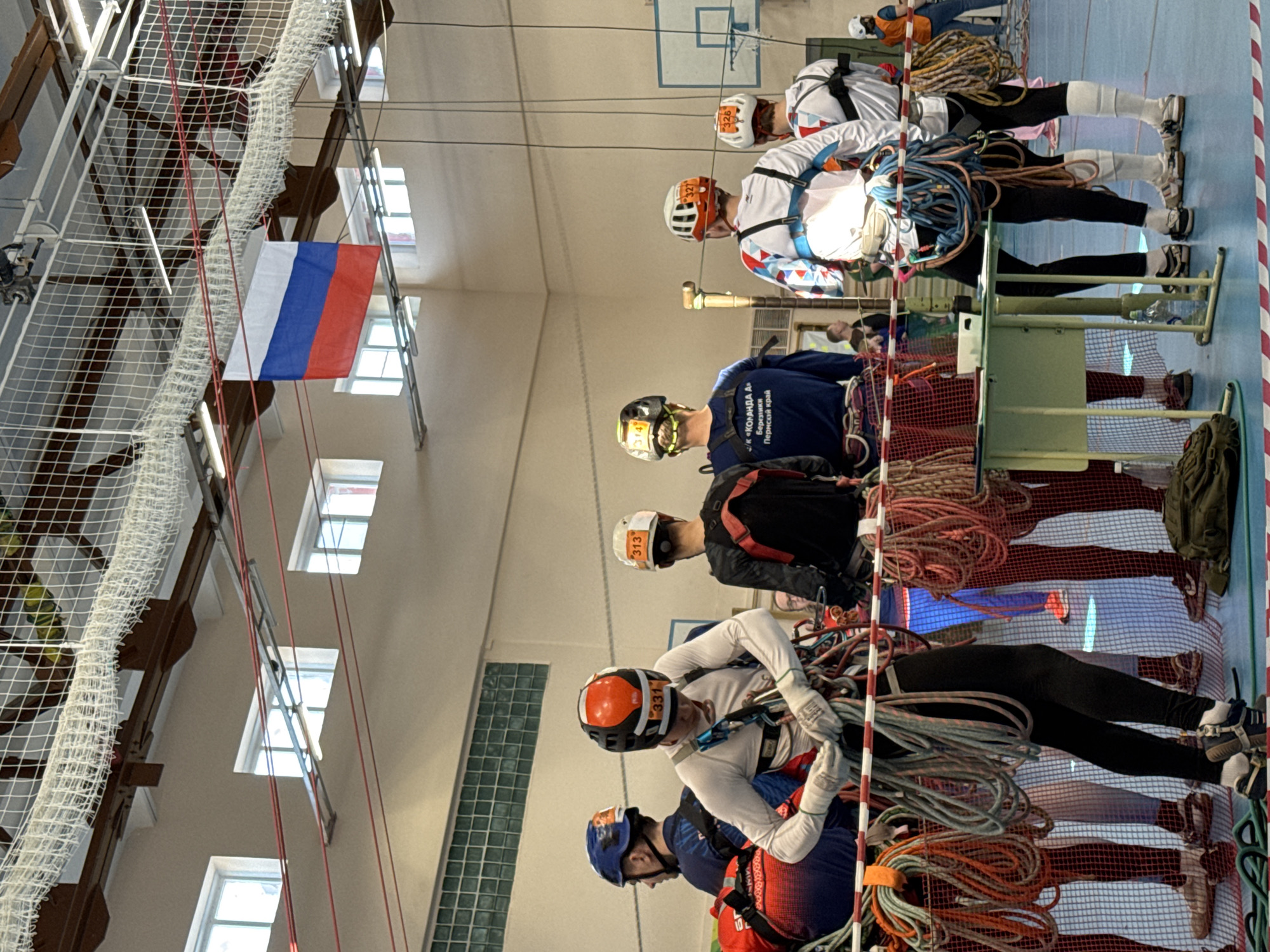 The image shows an indoor setting, possibly a gymnasium, with a group of people engaged in what appears to be a climbing or mountaineering exercise. Several individuals are wearing helmets and harnesses with ropes attached. They are positioned along a structure that might be a climbing wall or a setup for practicing rope techniques.
A Russian flag is visible in the upper left corner, which provides context and suggests a location within Russia. While the image does not directly specify Usolye, Russia, the mention of the city in the prompt indicates that this event likely takes place there. The environment includes elements of a gymnasium, like the basketball hoop in the background and the blue flooring with lane markings. The people are interacting with ropes, equipment, and each other, suggesting a training or competition scenario.