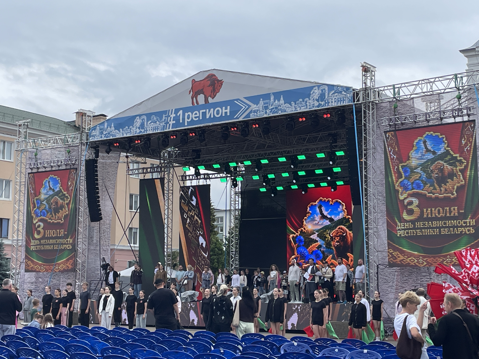 An outdoor event is taking place on a cloudy day in the city of Brest, Belarus. A large stage is set up with two large screens on either side displaying images. The screen on the left shows an eagle flying over a landscape with a bison. The screen on the right shows a similar landscape with a bison and text in Russian that reads "3 ИЮЛЯ - ДЕНЬ НЕЗАВИСИМОСТИ РЕСПУБЛИКИ БЕЛАРУСЬ," meaning "July 3 - Independence Day of the Republic of Belarus." Above the stage, a banner reads "#1 РЕГИОН" with an image of a bison. A crowd of people, including performers and spectators, are gathered in front of the stage, with rows of blue seats visible in the foreground. Some people on stage are dressed in black, possibly as performers. Other individuals in the audience appear to be engaged in conversation. Red and white Belarusian flags are visible. The buildings in the background are typical of urban architecture.