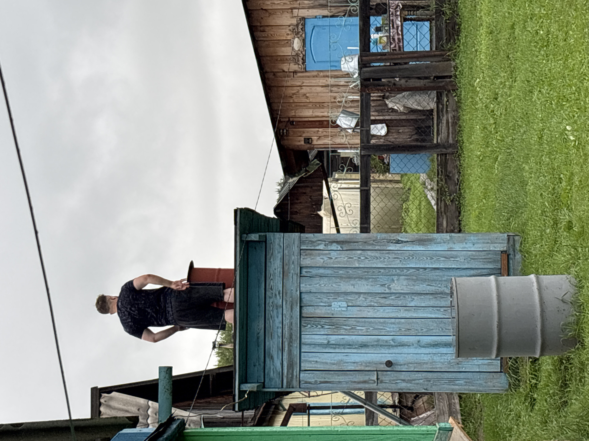 A man stands on top of a blue wooden outhouse in what appears to be a rural setting in the city of Gonja, Russia. The man, seen from the back, is wearing a dark t-shirt and dark shorts. He has his hands on his hips. The outhouse is weathered and has a hinged door on the front. A grey metal barrel sits on the grass in front of the outhouse. In the background, there is a wooden building with a blue door and a fenced yard containing several metal buckets. The sky is overcast.