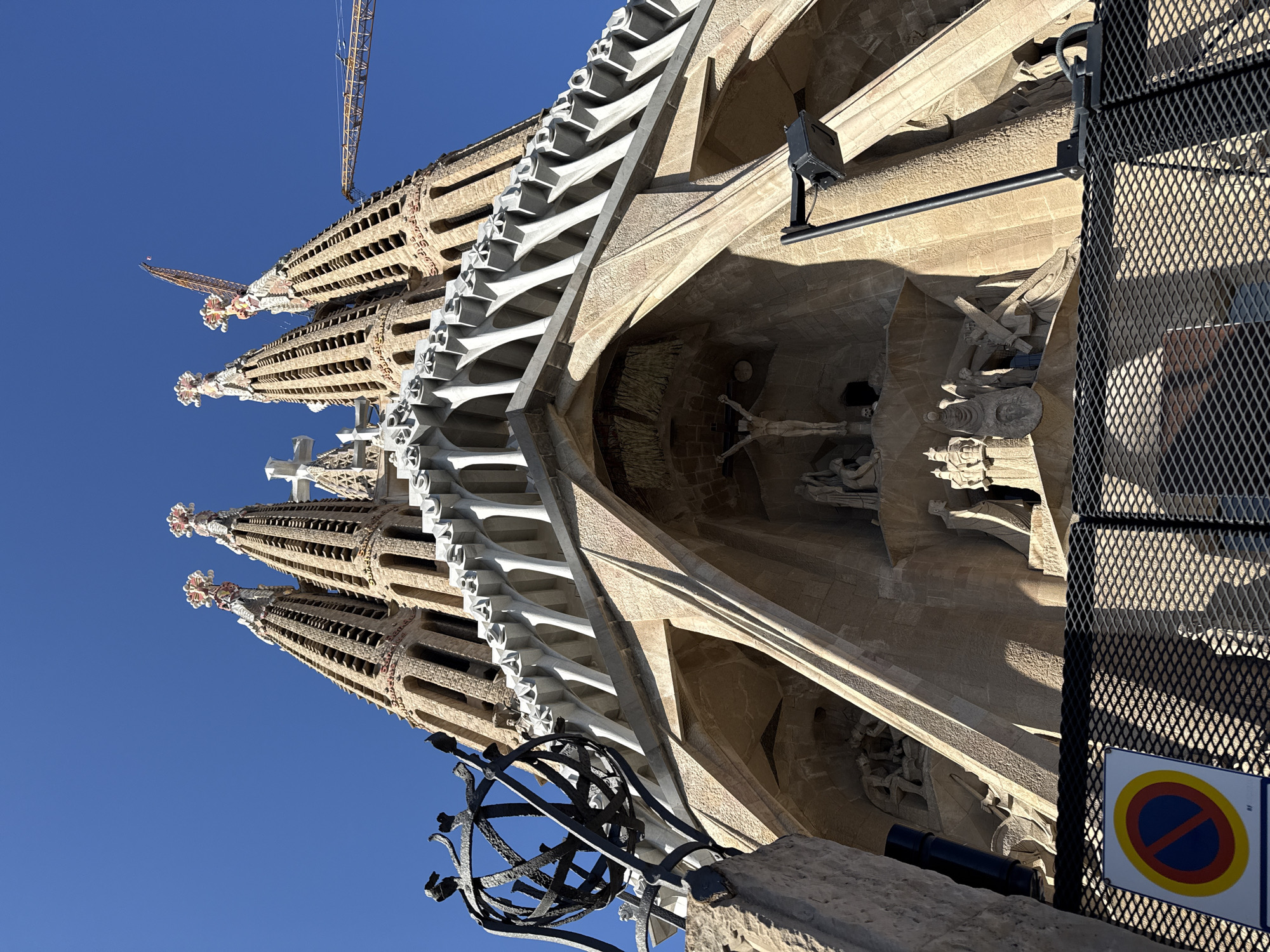 This image captures an upward-angled view of the Sagrada Família in Barcelona, Spain, specifically focusing on a portion of the Passion Façade under a clear blue sky. In the immediate foreground, a black metal mesh fence partially obscures a white circular sign with a red border, a blue inner circle, and a red diagonal line, indicating "no stopping." To the left, a dark, ornate, spherical metal structure with radiating spikes is prominently featured.
The main subject is the towering stone facade of the Sagrada Família. Its lower section features numerous sculpted figures in an angular, expressive style, depicting biblical scenes. A large crucifix hangs above a central archway, with other figures and architectural details visible around it. Strong sunlight illuminates the textured stone, casting deep shadows that highlight the intricate carvings and structural elements.
Above the main sculptural facade, a prominent horizontal band of white, geometric, angular architectural elements runs across the structure. Beyond this, the building ascends into multiple conical spires made of light-colored stone. Two prominent spires are visible in detail, each adorned with numerous apertures and capped with ornate, multi-colored finials. A white cross is visible on a spire further to the left. In the upper right quadrant of the image, the arm of a yellow construction crane extends into the sky, indicating ongoing work on the basilica. Another crane boom is visible extending upward behind the two prominent spires. The background consists of an unbroken, vibrant blue sky. No individuals are visible within the frame.