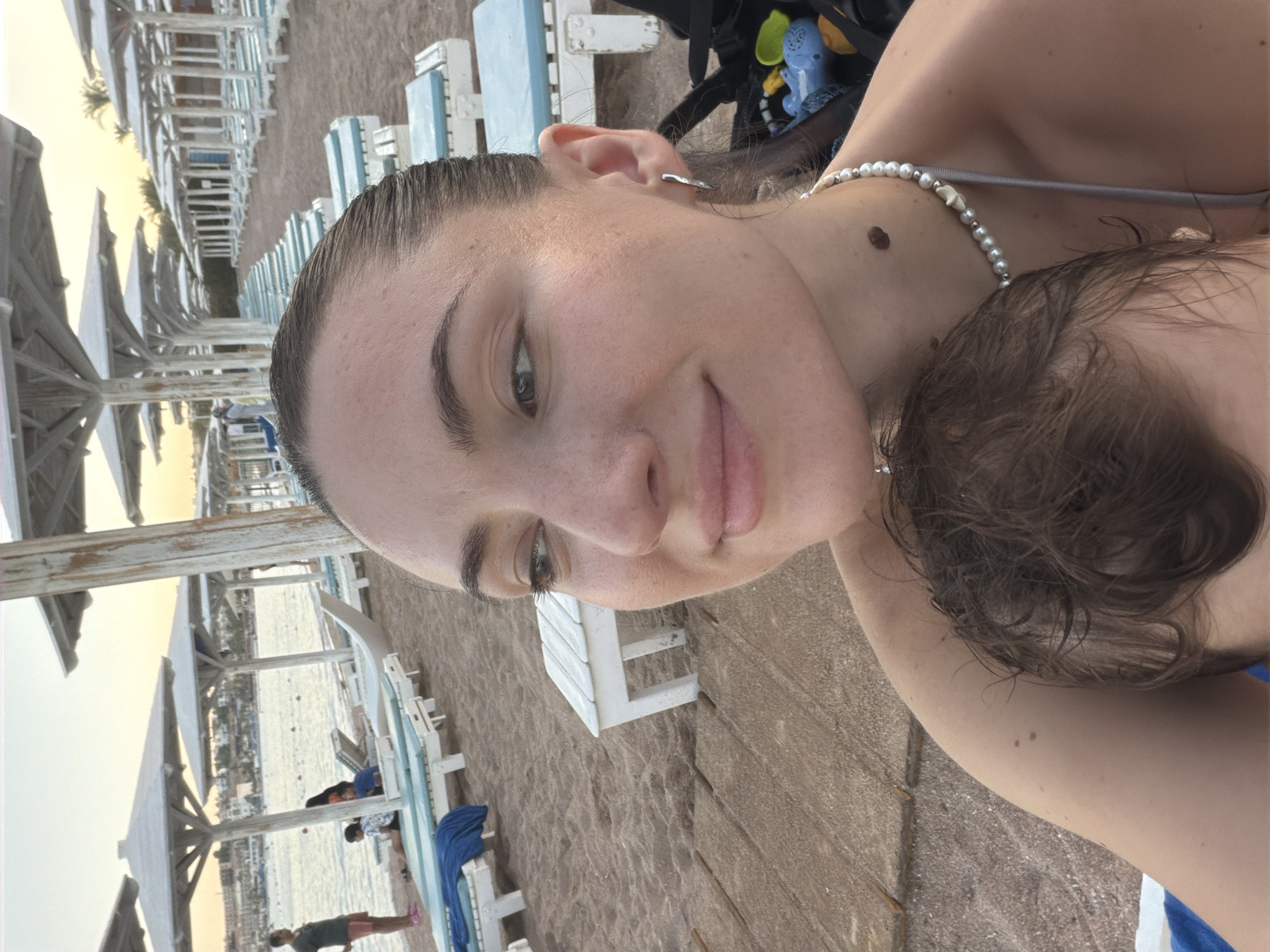This is a selfie taken at a beach in Sharm El Sheikh, Egypt. The foreground is dominated by the face of a young woman with her hair pulled back. She is looking to her left, and her expression is a slight smile. She is wearing a pearl necklace. Partially obscured by her is a young child's head, showing brown, wet hair.
The background of the image depicts a beach scene. Rows of empty white lounge chairs with blue cushions are lined up under white umbrellas. The sand is light brown, and further back, the sea is visible with a few small boats and buildings along the coastline. The sky is a soft orange-pink, suggesting it is either sunrise or sunset. The overall impression is a peaceful, relaxed atmosphere at a tourist resort.