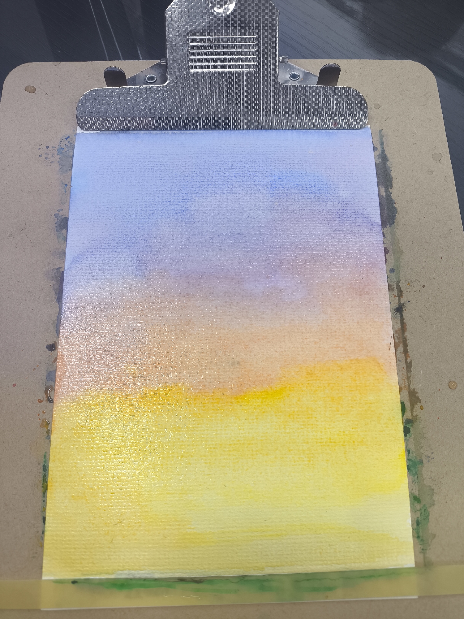 A watercolor painting, depicting a gradient of colors from yellow at the bottom, transitioning to orange in the middle, and then to blue at the top, is clipped to a clipboard. The clipboard is metallic at the top and composed of light brown cardboard below. A strip of tape, colored light green, is visible along the bottom edge of the paper. The clipboard rests on a dark surface, which is presumed to be a table or desk. There are no indications as to the location being Moscow, Russia. There are no people, actions, or interactions depicted.