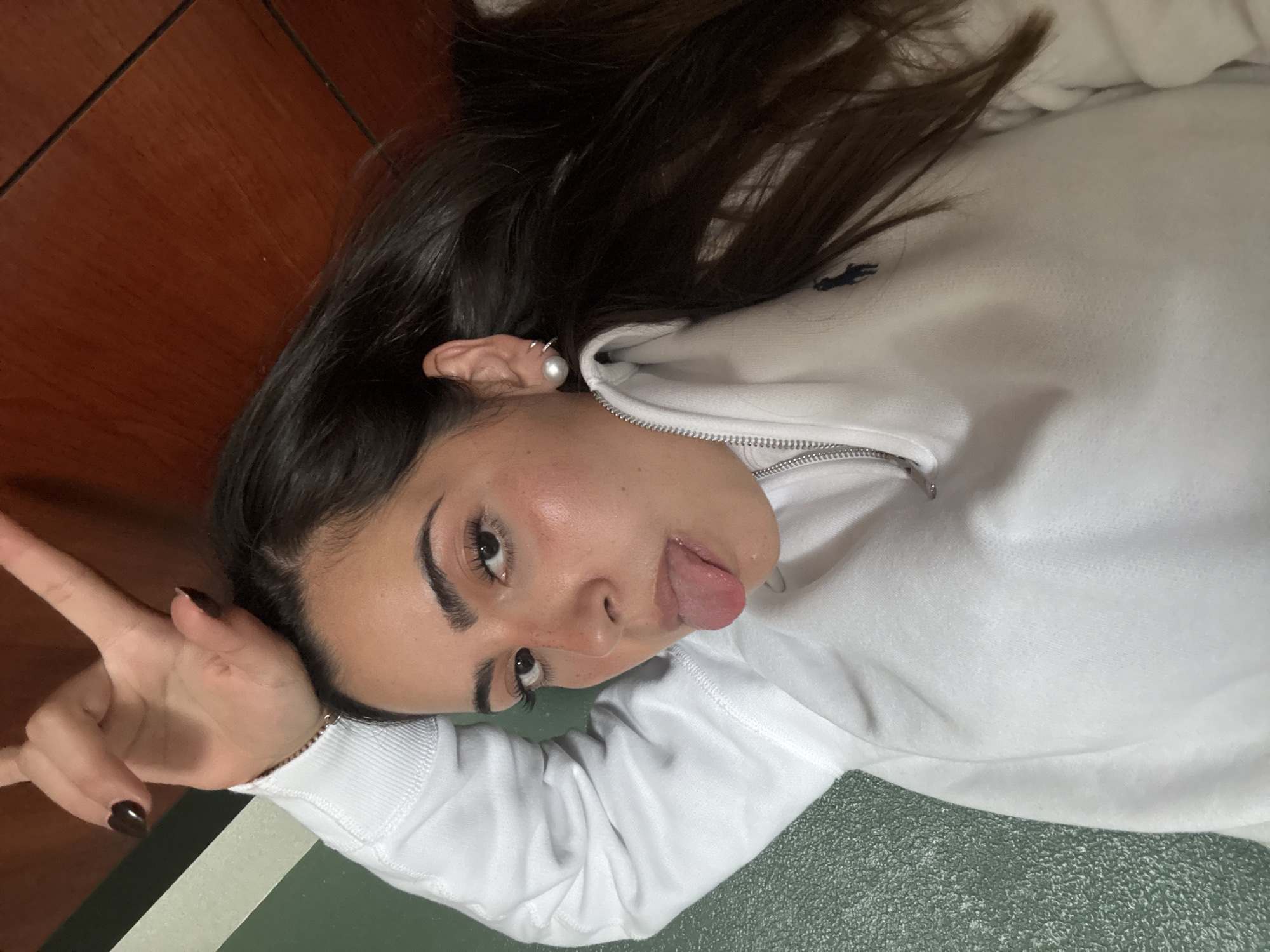 A young woman is lying on her back with her head tilted, sticking her tongue out. She is wearing a white zip-up jacket. Her hair is dark brown and long. She is wearing pearl earrings and has dark fingernail polish. She is making a peace sign with her left hand. The background consists of a wooden surface and a green and white surface. The location is San Antonio, United States of America.