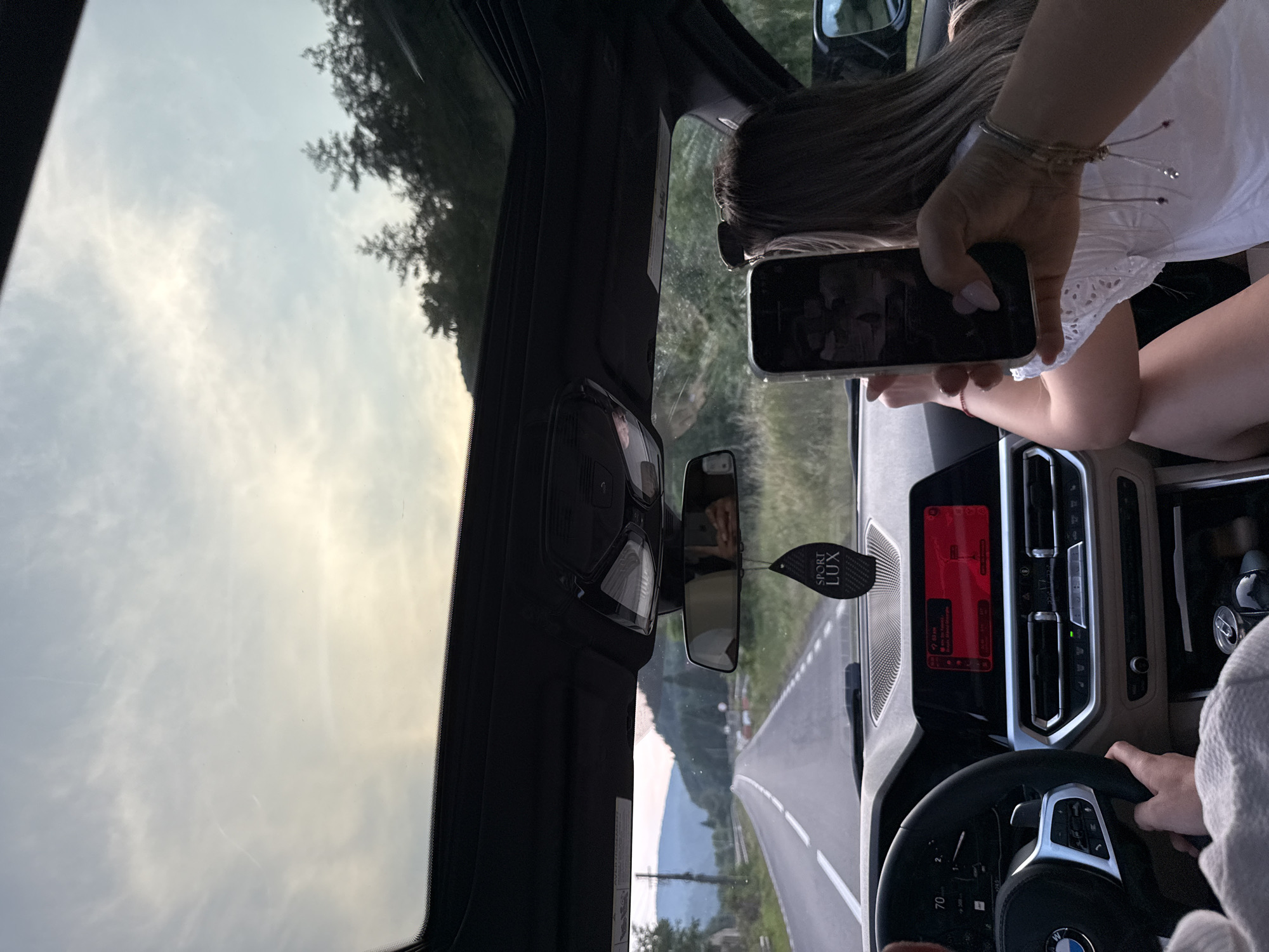 The image captures an interior view of a car traveling on a road in or near Călcâi, Romania, likely during sunset or early evening given the lighting. The perspective is from the passenger seat, looking forward through the windshield and upwards through the sunroof.

In the foreground, a person's hand holds up a smartphone, taking a picture or video. The phone is angled towards the front of the car and the landscape outside. A woman with long, blonde hair, wearing a white top, is seated in the passenger seat, with her back mostly to the camera. Her arm and shoulder are visible as she holds the phone.

The car's interior is visible, including the dashboard with its digital display showing navigation and other vehicle information, the steering wheel held by another person (partially visible on the left), and air vents. A "SPORT LUX" air freshener hangs from the rearview mirror.

Outside the car, a paved road stretches into the distance, bordered by fields and trees on either side. Rolling hills and mountains are visible in the background, with a hazy sky suggesting dusk. The road appears to be a rural route. The presence of a road and natural landscape is consistent with the rural setting of Călcâi, Romania.