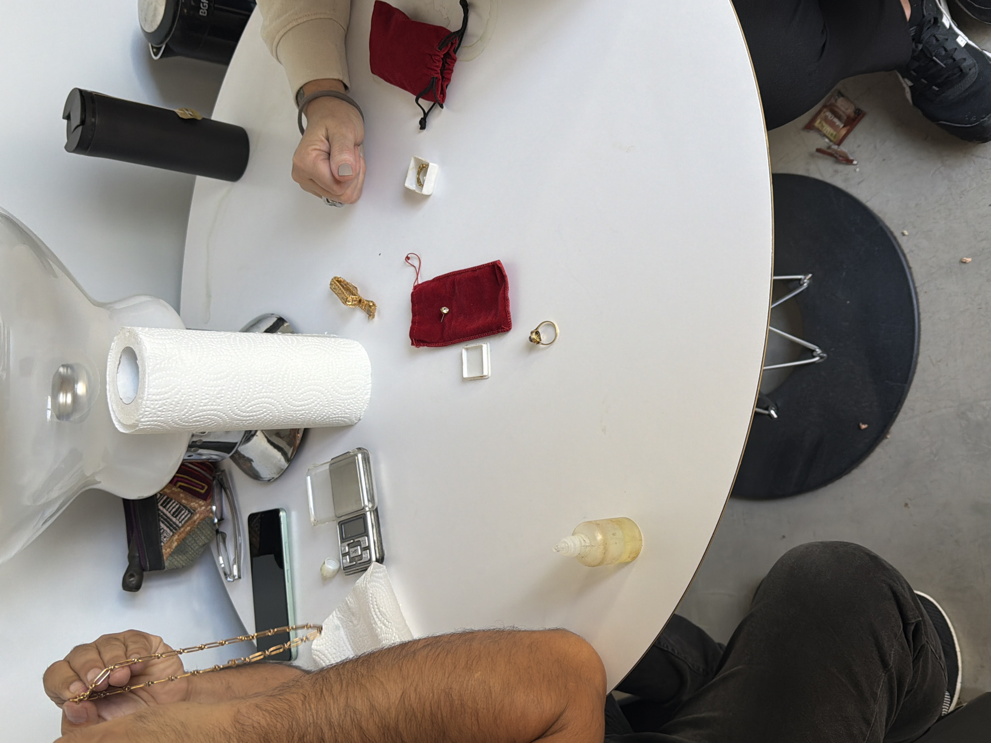 A high-angle, slightly rotated photograph depicts a white, round table surface with various items scattered across it. The view is from above, capturing portions of two seated individuals along the table's edge.
To the upper left, a portion of a person's arm and hand wearing a bracelet are visible near a red velvet drawstring bag, a small white box, and a gold brooch. A black cylindrical object is also in the background.
Center table includes another red velvet drawstring bag, a gold ring, a small white box, a partially used roll of paper towels, a mobile phone, a scale, and a bottle with a yellow liquid.
A person holding a gold chain and a partially obscured brown arm and hand are visible.
To the right of the table are lower legs of seated individuals wearing dark-colored trousers and sneakers and a black cushion.
The location cannot be determined based solely on the visual information provided in the photograph.