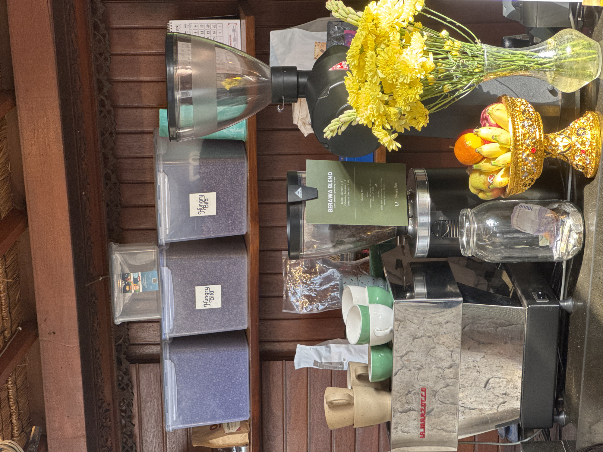 The image depicts a coffee shop counter at Hungry Bird Coffee in North Kuta, Indonesia. In the foreground, a La Marzocco espresso machine sits to the left. Above the machine, several coffee cups are stacked. To the right of the espresso machine, a glass jar filled with coins is visible. Further to the right, a vase of bright yellow flowers stands next to a golden fruit bowl filled with bananas, apples, and an orange. Behind the fruit bowl, a coffee grinder with a large bean hopper is present. On a shelf above, several clear plastic containers hold coffee beans, labeled with the Hungry Bird logo. A calendar is also visible on the shelf. The counter is made of dark material, and the background features wooden shelving and panels. The overall setting suggests a typical coffee shop environment with a focus on coffee preparation and presentation. There are no people visible in the image.