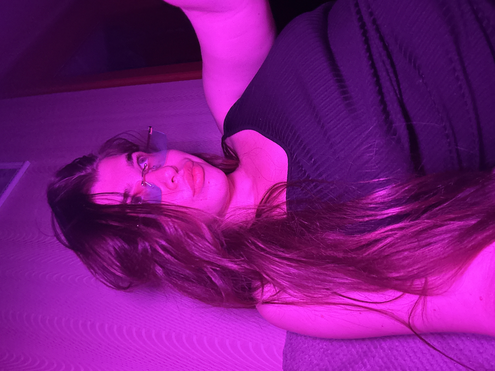 A young woman is seated, leaning back on a couch with a textured gray upholstery. She is wearing a black ribbed tank top and light-colored, possibly blue-tinted, sunglasses with thin metal frames. Her long, dark brown hair is down and spread across her shoulders and back, obscuring part of her body. Her face is turned slightly to her left, with her lips appearing plump and slightly parted. The lighting in the scene is predominantly a vibrant magenta or pink hue, casting a strong colored light on her skin, hair, and the surrounding environment. The background consists of a wall with a subtle, wavy texture. To the right of the woman, there is a darker area, possibly a doorway or furniture, with a vertical reddish-brown element visible. The overall impression is that of a selfie or a casually taken photograph, likely indoors, with the artificial lighting creating a dramatic effect. No specific location in Мещёры, Russia, can be determined from the visual information provided.