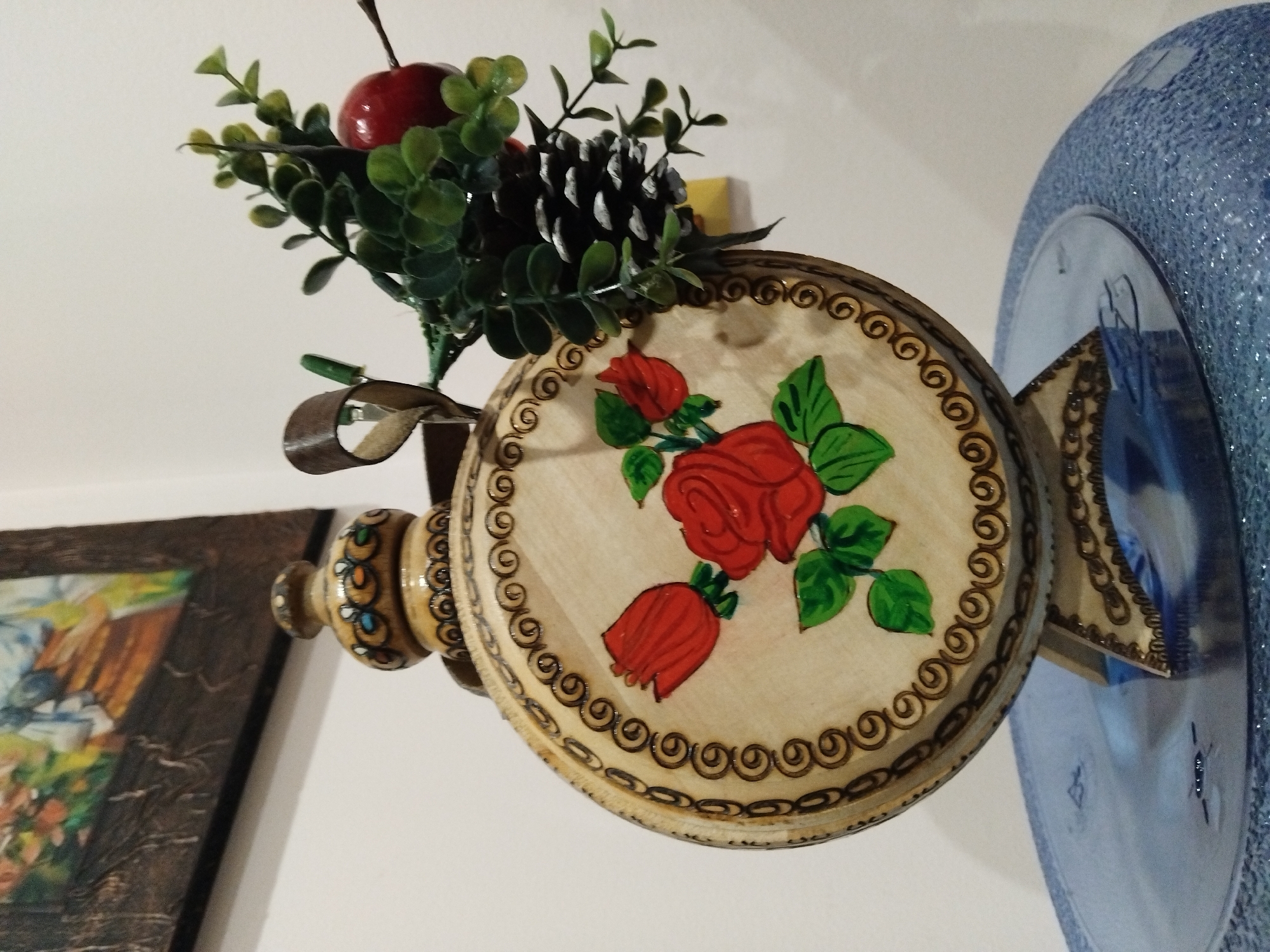 A still life shot is taken indoors. The focus is a decorative wall hanging with a wooden plate featuring painted red roses and green leaves against a light background, bordered by a swirling pattern. A cluster of artificial greenery, including a red apple and a pinecone, is attached above the plate. The wall hanging is embellished with carved wooden elements. In the background, there is a framed painting, partially visible on the left, and a blue, textured decorative object on the right. The location is Nova Zagora, Bulgaria, suggested by the presence of cultural artifacts or decorations typical of the region. The interaction between objects in the scene is purely compositional.