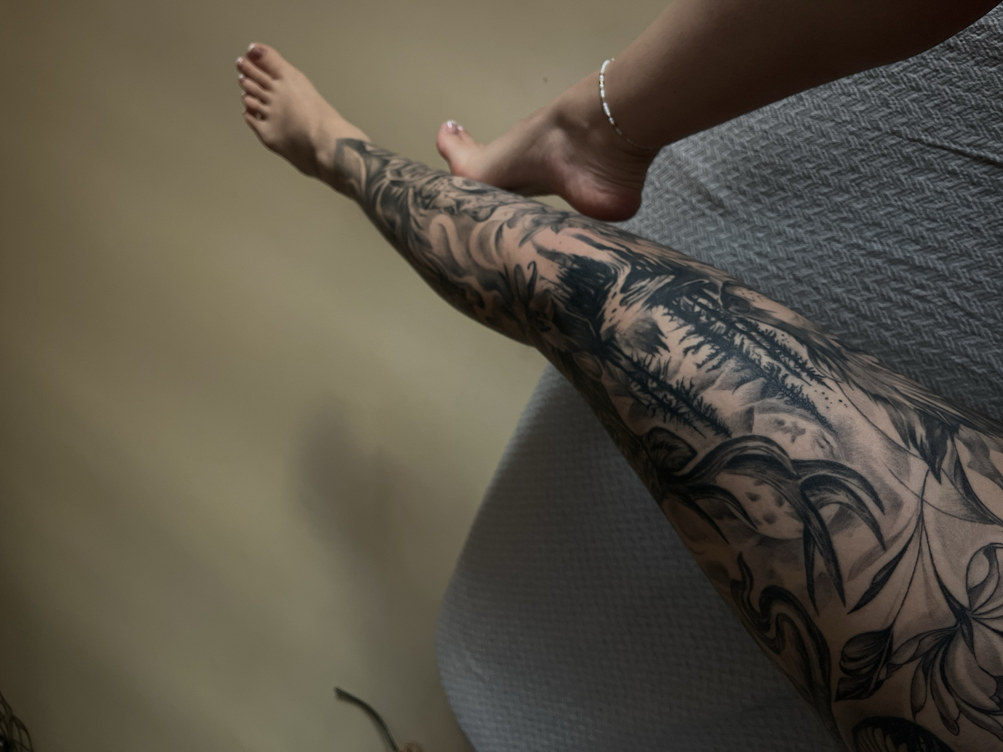 The image shows a close-up view of a woman's legs with extensive black and grey tattoos covering her left leg from ankle to thigh. The tattoos depict a forest scene with trees, mountains, and possibly clouds or smoke. Her right leg is visible from the knee down, with her foot resting near the tattooed leg. She is wearing a delicate silver anklet on her right ankle. The woman appears to be reclining on a textured gray surface, possibly a bed or sofa. The background is a plain, neutral-colored wall. The lighting suggests an indoor setting. The specific location in Veliko Tarnovo, Bulgaria, is not identifiable from the image.