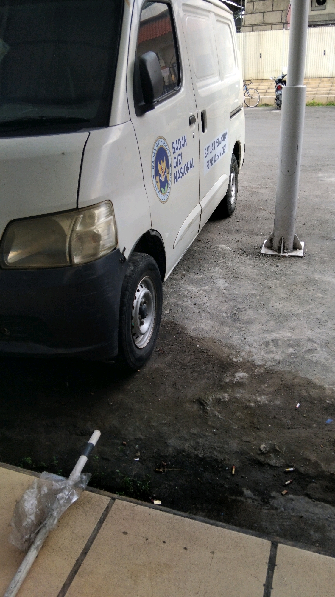 The image shows the side of a white van with the text "BADAN GIZI NASIONAL" and "SATUAN PELAYANAN PEMENUHAN GIZI" visible on its side. The van is parked on a paved surface that appears to be a street or lot, with some dirt and debris on the ground. In the background, a bicycle and a motorcycle are parked. A white pole is also present in the foreground to the right of the van. A broken white pole covered in plastic is on the left side of the image, lying on a tan tiled surface. The scene appears to be outdoors, possibly in an urban or semi-urban environment, with buildings visible in the background. There are no people clearly visible in the image. The overall impression is that of a parked vehicle in a public area.