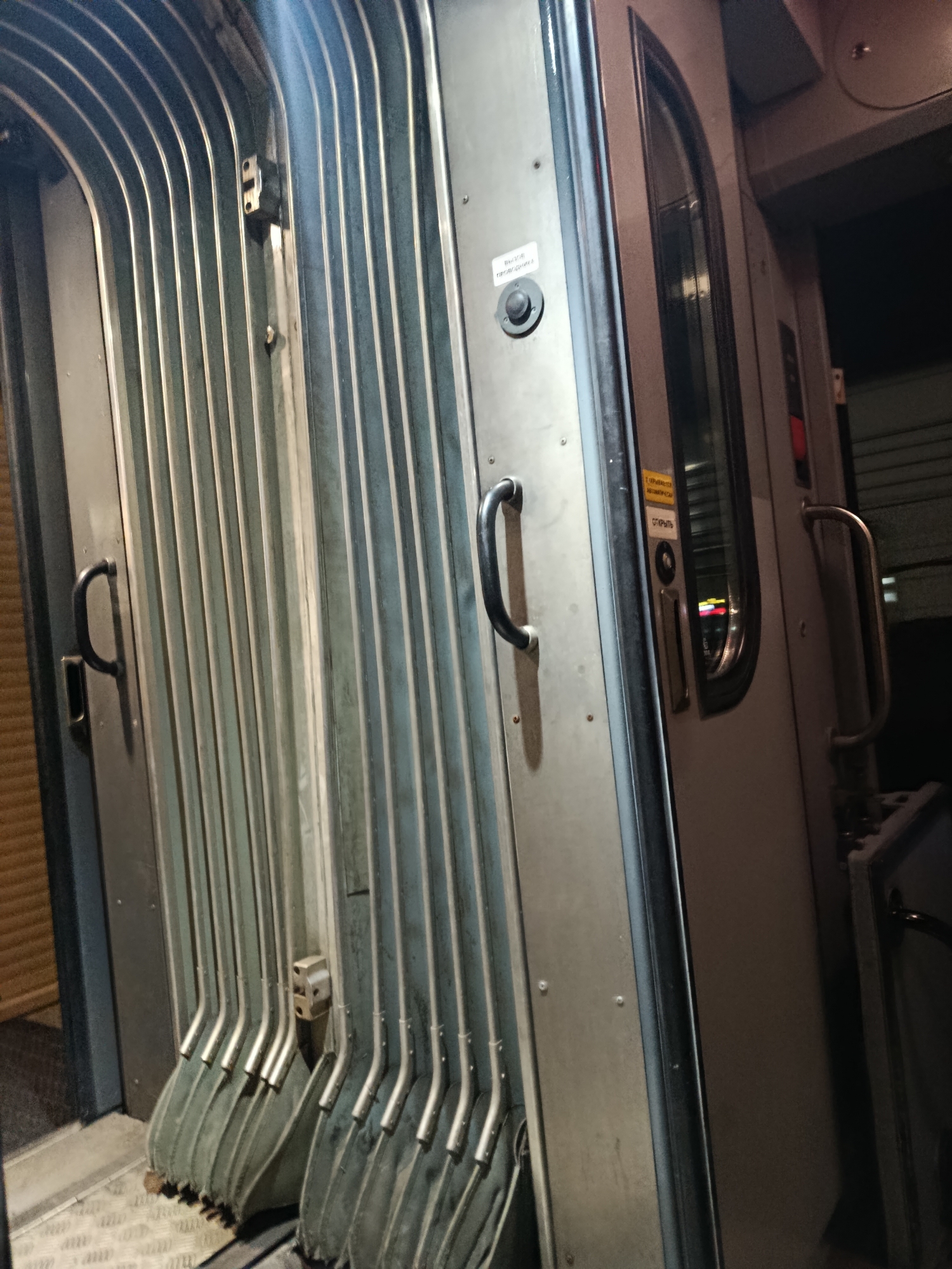 The image shows the exterior of a train car, likely at night. The focus is on a set of accordion-style doors that are partially open. The doors are made of ribbed metal panels and have black handles. To the right of the doors, there is a standard door with a window and a metal handrail. There are also some signs on the train car, but they are difficult to read. The ground in front of the train is a textured metal surface. The overall impression is of an industrial or transportation setting. The location in the city of Rtishchevo, Russia is not discernible from the image.