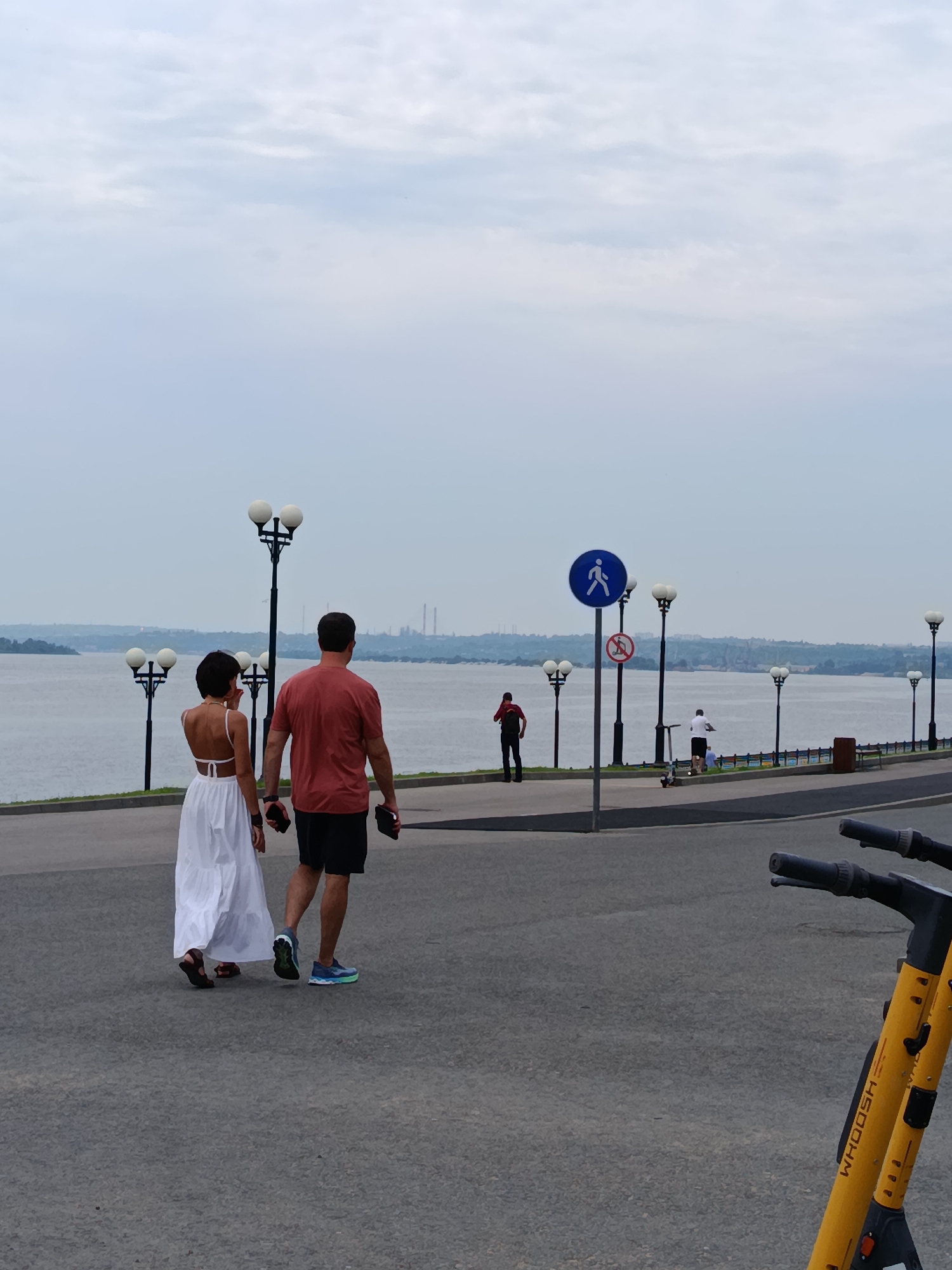 The image depicts a waterfront promenade in Saratov, Russia. A man and a woman are walking away from the camera, the woman in a white dress and the man in a red t-shirt and shorts. In the background, a wide river is visible, with industrial structures and a hazy skyline on the opposite bank. Several lampposts line the promenade, and signs indicate pedestrian and no-scooter zones. Other individuals are seen in the distance, some on scooters. In the foreground, a yellow electric scooter is partially visible. The sky is overcast, suggesting a cloudy day.