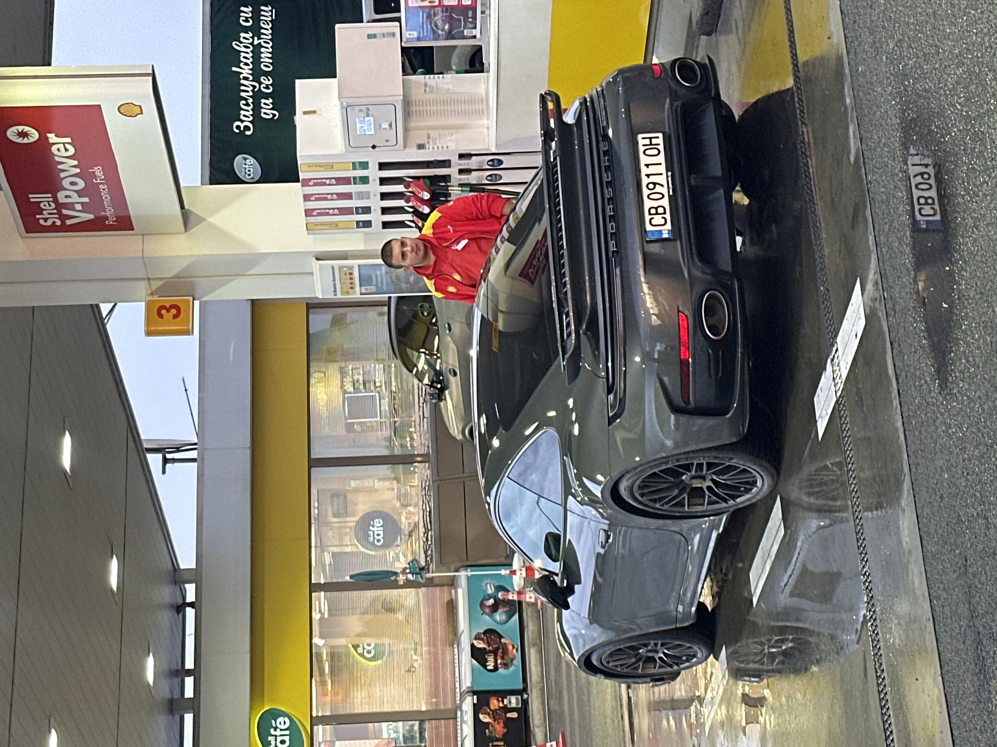 A dark grey Porsche 911 GT3 is parked at a Shell V-Power gas station in Drazhevo, Bulgaria. The car is positioned at a fueling pump, and its rear is facing the camera. The license plate reads "CB 0911 OH". A gas station attendant wearing a red and yellow uniform stands near the pump, looking towards the car. The ground is wet, reflecting the car and the surrounding lights, suggesting recent rain or cleaning. In the background, the Shell V-Power signage is prominent, along with other gas station amenities and shop windows displaying food items. The overall scene depicts a routine refueling stop at a modern gas station.