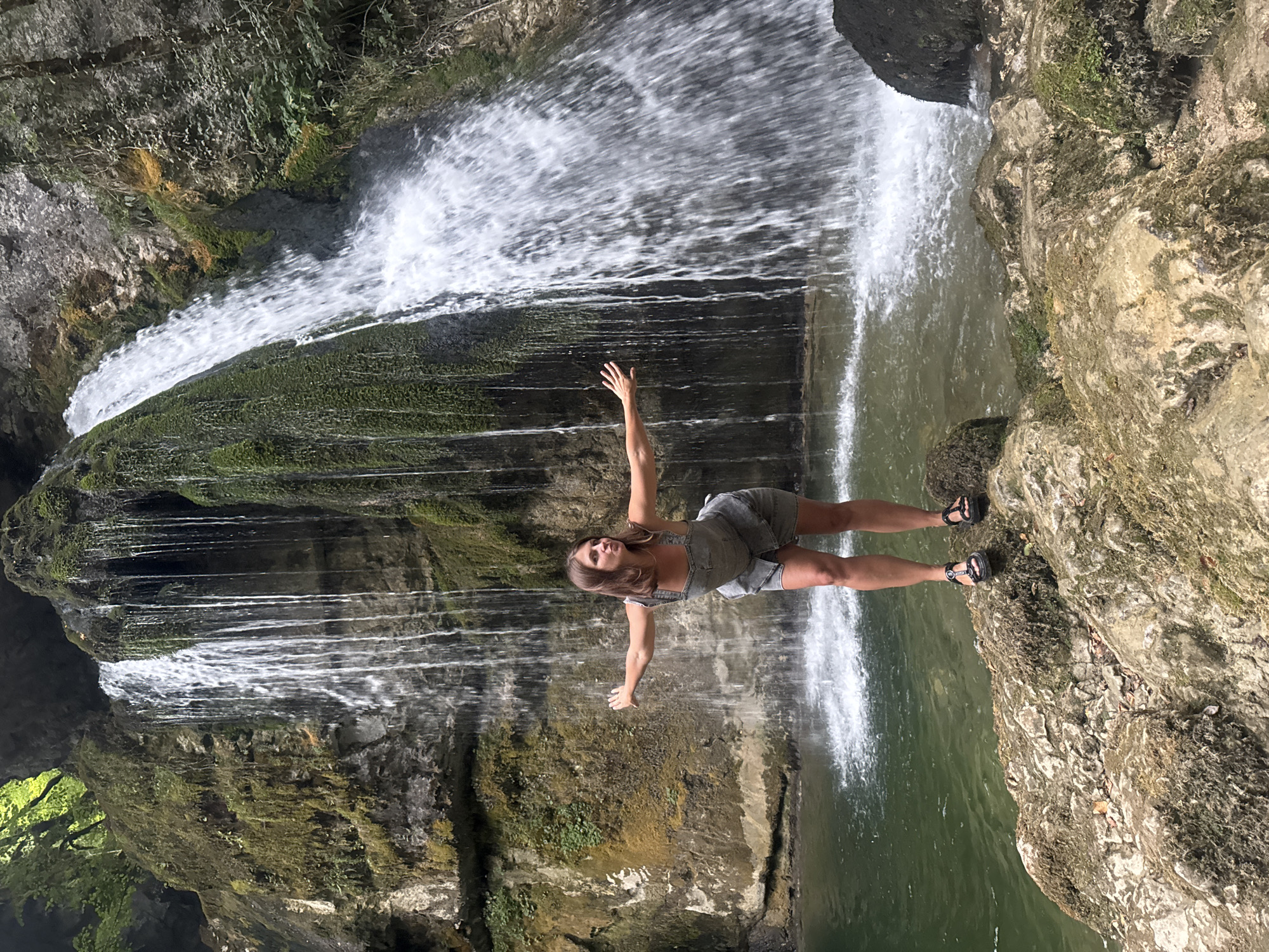 A young woman with brown hair, wearing a denim dress and sandals, stands on moss-covered rocks in front of a waterfall in the Jura Mountains near Saint-Claude, France. The waterfall cascades down a rocky, moss-covered cliff face into a clear green pool of water. The woman has her arms outstretched, her head tilted back, and her mouth slightly open as she looks off to the side. The scene is natural and lush, with green foliage visible at the top of the frame.