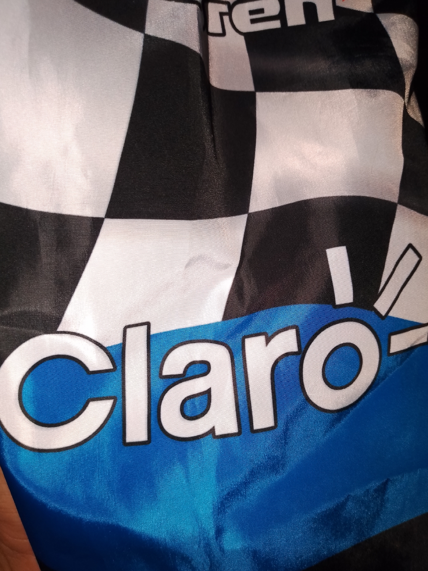 The scene, located in the city of Santa Ana, Venezuela, displays a piece of synthetic fabric, likely a flag or banner. The fabric features a black and white checkerboard pattern in its upper portion, characteristic of a racing checkered flag. Below this, a solid blue section is present. Prominently displayed in white, sans-serif letters with a black outline, the word "Claro" is centered across the fabric, overlapping from the checkerboard into the blue area. Partially visible white letters spelling "ren" are situated above the "Claro" logo. The fabric exhibits fine horizontal striations and a slight sheen, suggesting a synthetic material, and is creased in several places. A small portion of a human hand is visible in the bottom-left corner, interacting with the fabric. The illumination of the scene is uneven, casting highlights and shadows across the material.