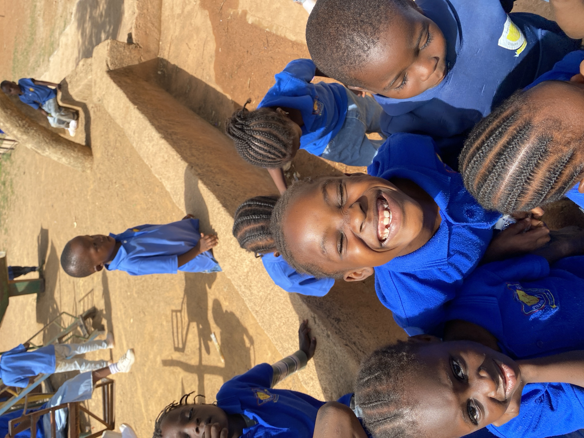 The image shows a group of children wearing blue uniforms and gathered outside. One child is laughing and looking up toward the camera. Other children are around them, some with braided hair. In the background, there's playground equipment and an area that looks like concrete structures or a building. The children are wearing blue uniforms with an emblem that reads "Rayfield". Based on the emblem and the location metadata, it is likely the photograph was taken at or near Rayfield in Gura Top, Nigeria.