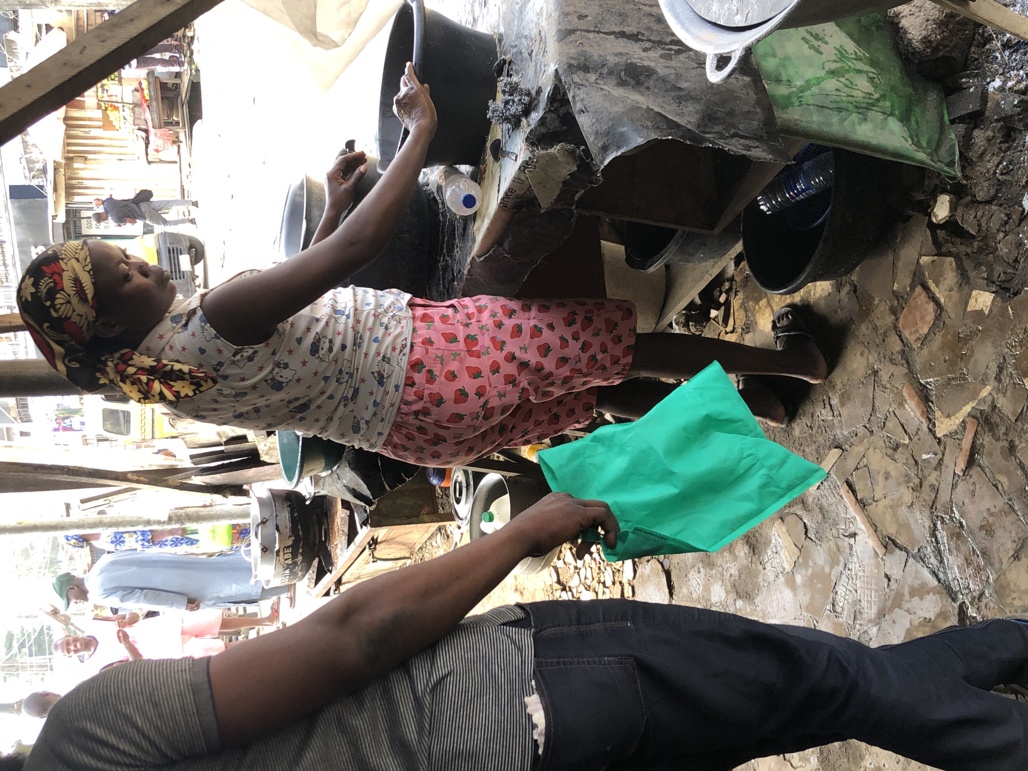 The image captures a scene in an outdoor market, possibly in Ikeja, Nigeria. A person, identified as a woman based on her attire, is the central focus, wearing a headscarf and a light-colored top with a strawberry print. She is positioned behind a makeshift counter, appearing to be engaged in some activity involving large black basins. Her right hand is submerged in one of the basins, suggesting she might be washing something. Another person, partially visible on the left, is wearing a striped shirt and dark pants, holding a bright green plastic bag. The environment appears somewhat disheveled, with broken tiles on the ground and various containers and objects scattered around. In the background, other people and market stalls are visible, indicating a bustling commercial area. The overall impression is of everyday market life, with elements of commerce and activity.
