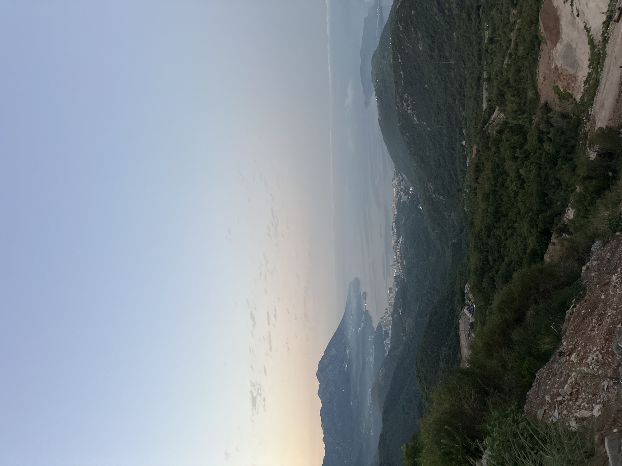 This is a landscape photograph taken from a high vantage point overlooking a coastal area at dusk. The sky is a pale blue with a gradient towards a light pinkish hue near the horizon, suggesting the sun has recently set or is about to rise. Wispy clouds are scattered across the sky.
In the middle ground, a bay curves into the land, with a town or city nestled along its shores. The buildings are illuminated with numerous lights, indicating it is twilight or early evening. Mountains, covered in dense green vegetation, rise from the land and frame the bay. Some of these mountains are covered in a light mist or haze, particularly those further in the distance.
In the foreground, the terrain is rough and rocky, with some sparse vegetation. There is evidence of construction or excavation, with piles of dirt and gravel visible. The overall scene depicts a natural landscape meeting a developed urban area, with the atmospheric conditions of dawn or dusk adding a serene and slightly muted quality to the image. The location is identified as being near Lapčići, Montenegro.