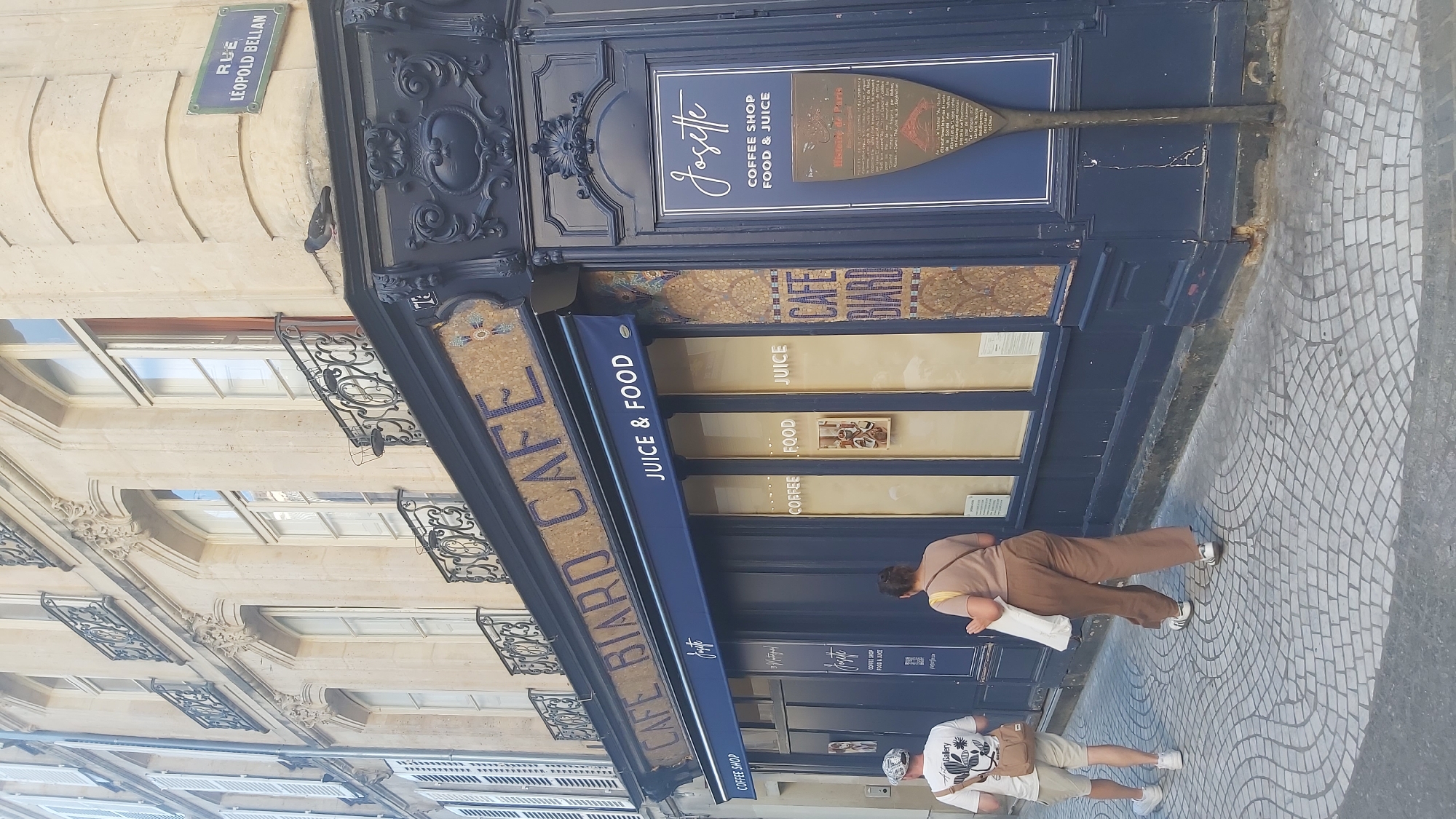The image depicts the exterior of a cafe called "Cafe Biard" located in Paris, France, as indicated by the street sign "RUE LEOPOLD BELLAN". The cafe has a dark blue facade with ornate decorative elements and mosaic detailing. A blue awning with "JUICE & FOOD" written on it is extended over the entrance. A historical plaque is mounted on the right side of the building. Two individuals are walking away from the camera on a cobblestone sidewalk. The person on the left is wearing a white t-shirt and shorts, and the person on the right is wearing a light brown top and pants. The building's architecture suggests it is in an older part of the city, with classical design elements visible on the upper floors, including balconies with wrought-iron railings and arched windows.
