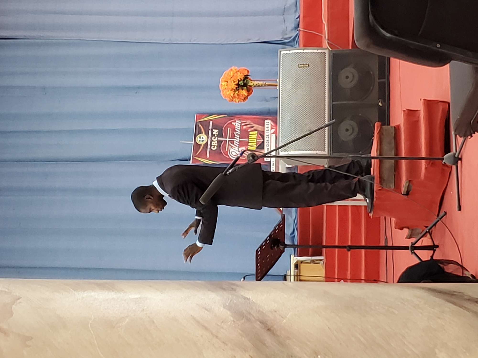 The image depicts a man standing on a stage in Unknown, Unknown. He is wearing a dark suit with a white shirt and is looking down with his hands extended. Behind him, there is a light blue curtain, a speaker, a sign with the words "CRC-N," "Mtumishi," and "Kauna," orange flowers, and red stairs. There is also a music stand and a microphone stand.