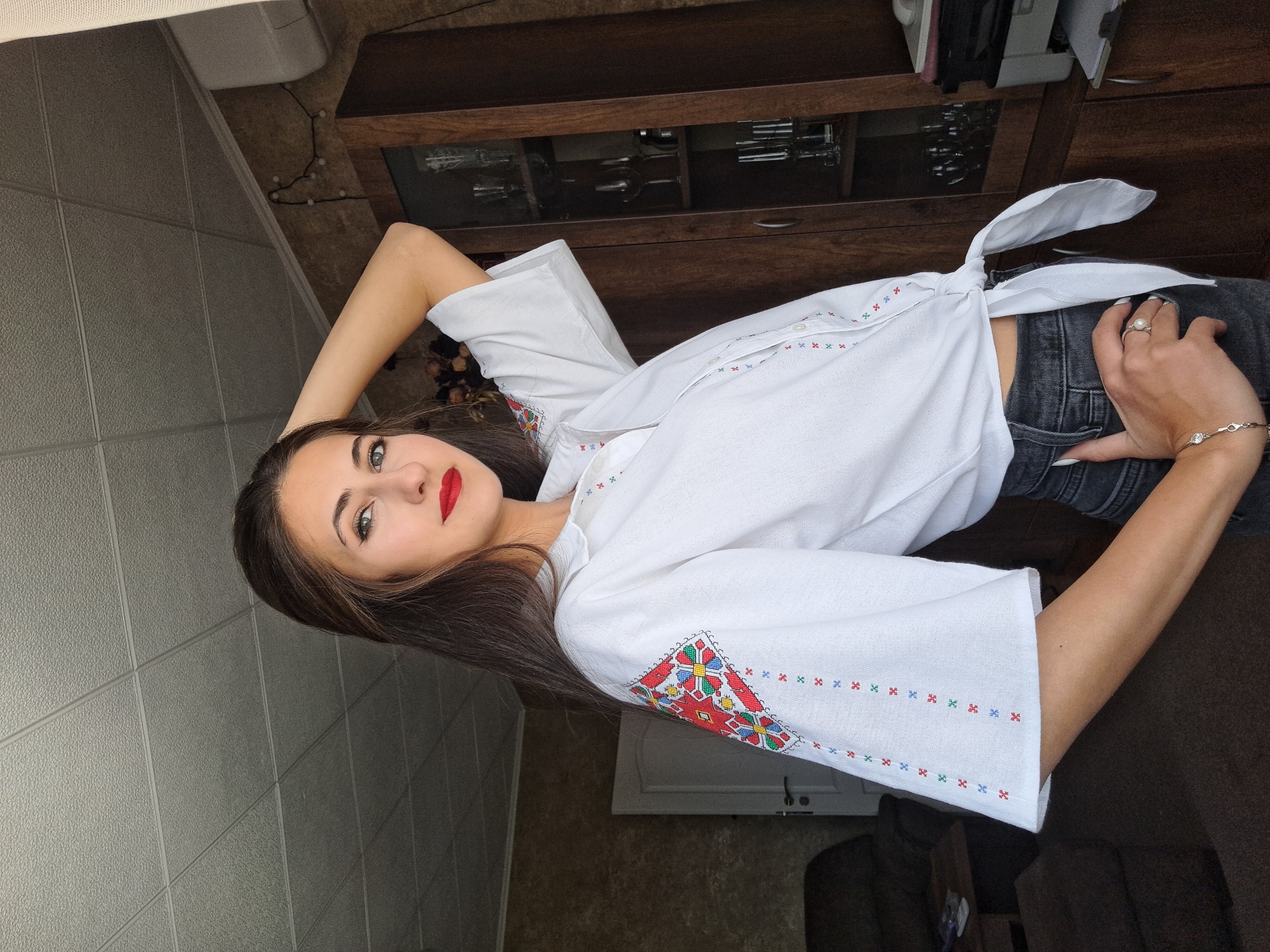 A woman is photographed indoors. She is wearing a white, short-sleeved button-up shirt with embroidery on the sleeves and collar, knotted at the waist. She is wearing grey jeans. Her left arm is raised behind her head and her right hand is on her waist. She has long brown hair and is wearing red lipstick. The background includes a dark wood cabinet, a tiled wall, and a brown wall. The location is Unknown, Unknown.
