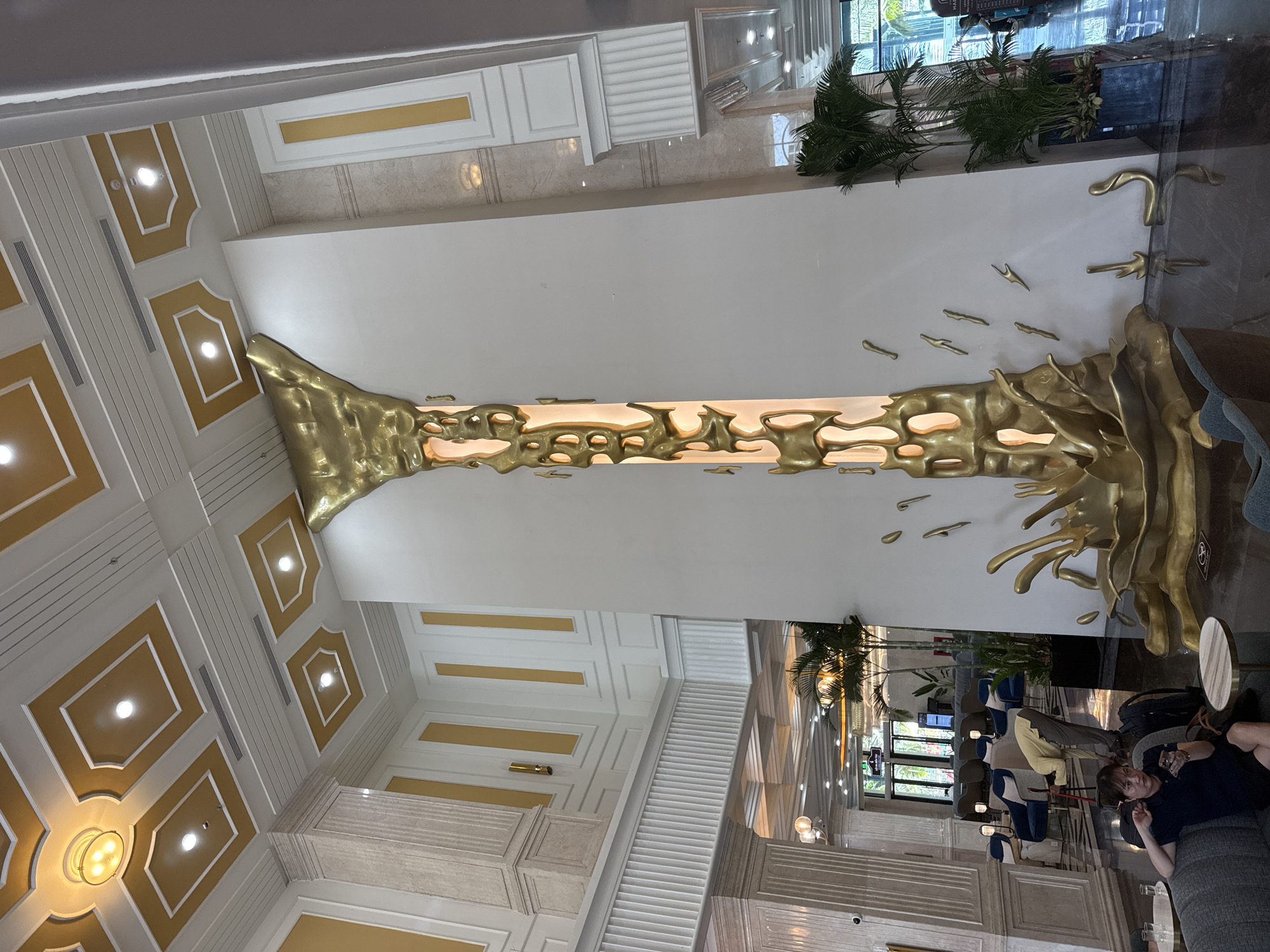 The photograph depicts the interior of a building in Nha Trang Ward, Vietnam. The ceiling is ornately decorated with recessed square panels outlined in gold. A large, golden, tree-like sculpture runs vertically along a white wall, spanning a significant portion of the wall's height.
Towards the right side of the image, a person is reclining on a grey sofa. The building interior includes various pieces of furniture, including chairs. Plants are situated near windows, suggesting a well-lit space with natural light.