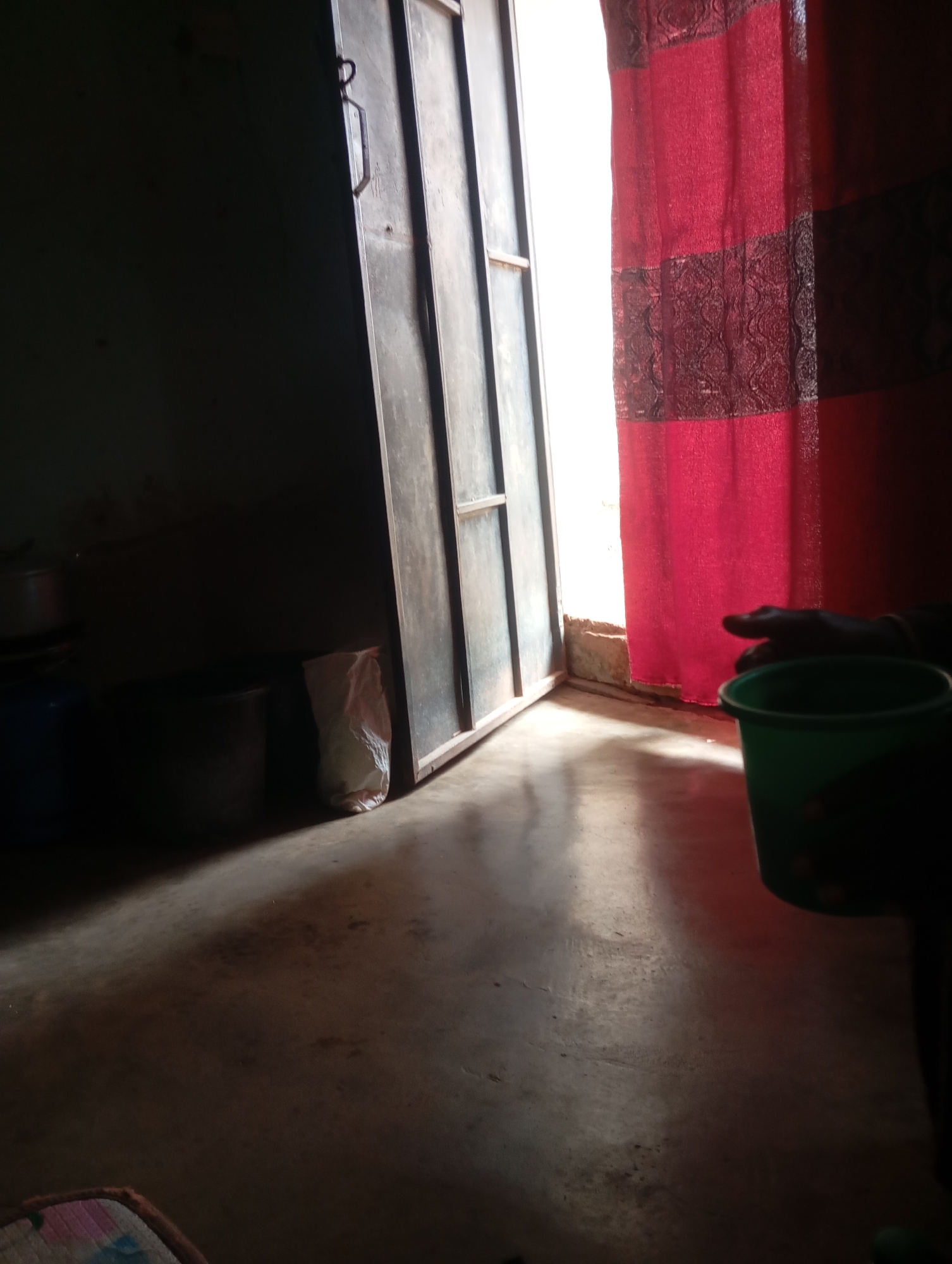 The image depicts an interior scene, likely a room or dwelling, with a focus on a doorway and a red curtain. Light streams in from the right, illuminating the lower portion of a metallic door frame and a section of the floor. The door itself is partially ajar, revealing a dark interior. To the right of the door, a bright red curtain with patterned horizontal bands hangs, suggesting a window or opening is present behind it.
In the foreground, a green plastic bucket is visible on the right side of the image, partially obscured by darkness. A hand is also present, reaching towards or interacting with the bucket. On the floor, in the mid-ground and to the left, several dark objects are present, including what appear to be large containers or pots, one of which has a light-colored bag next to it. The floor is concrete and shows some dust and wear. The overall lighting suggests it is daytime, with strong contrast between the brightly lit areas and the shadowed sections. The scene is consistent with a residential setting in Jalingo, Nigeria, characterized by the use of such materials and ambient lighting conditions.