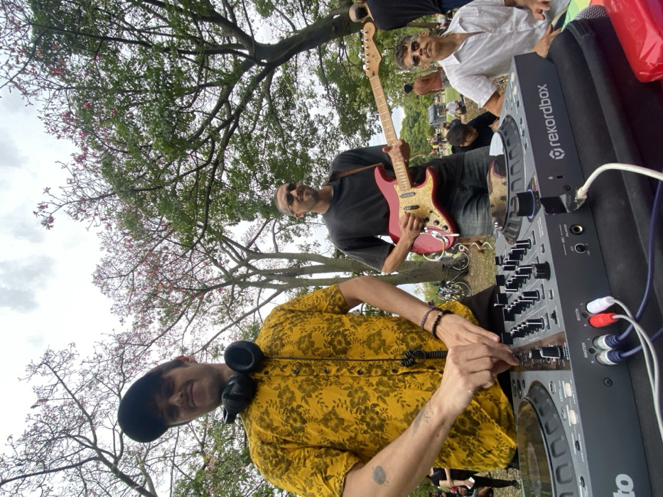 The image portrays an outdoor musical gathering in an urban park in Buenos Aires, Argentina, under an overcast sky. In the foreground, a light-skinned male, wearing a dark baseball cap and a yellow short-sleeved shirt with a dark floral pattern, operates a grey Pioneer DJ Rekordbox branded controller. Black over-ear headphones rest around his neck. His hands are positioned over the mixer section of the controller, manipulating faders and knobs. A circular tattoo is visible on his left forearm, and bracelets adorn his right wrist. To the right of the DJ, a second light-skinned male with short dark hair and sunglasses, dressed in a dark grey t-shirt and blue jeans, holds and appears to play a red Fender Stratocaster-style electric guitar featuring a light maple neck and a pearloid pickguard. A brown strap supports the guitar. To the right of the guitarist, a third light-skinned male, seated on the ground, wearing a white short-sleeved shirt and sunglasses, with a distinct mustache, observes the activity. The background consists of several large trees, some with green leaves and others exhibiting sparse pink blossoms. A sparse crowd of blurred individuals is visible scattered across the green grass in the mid-ground and background, and multiple bicycles are parked in the mid-ground. The DJ controller is connected by white, red, and blue RCA cables, and white TRS cables.