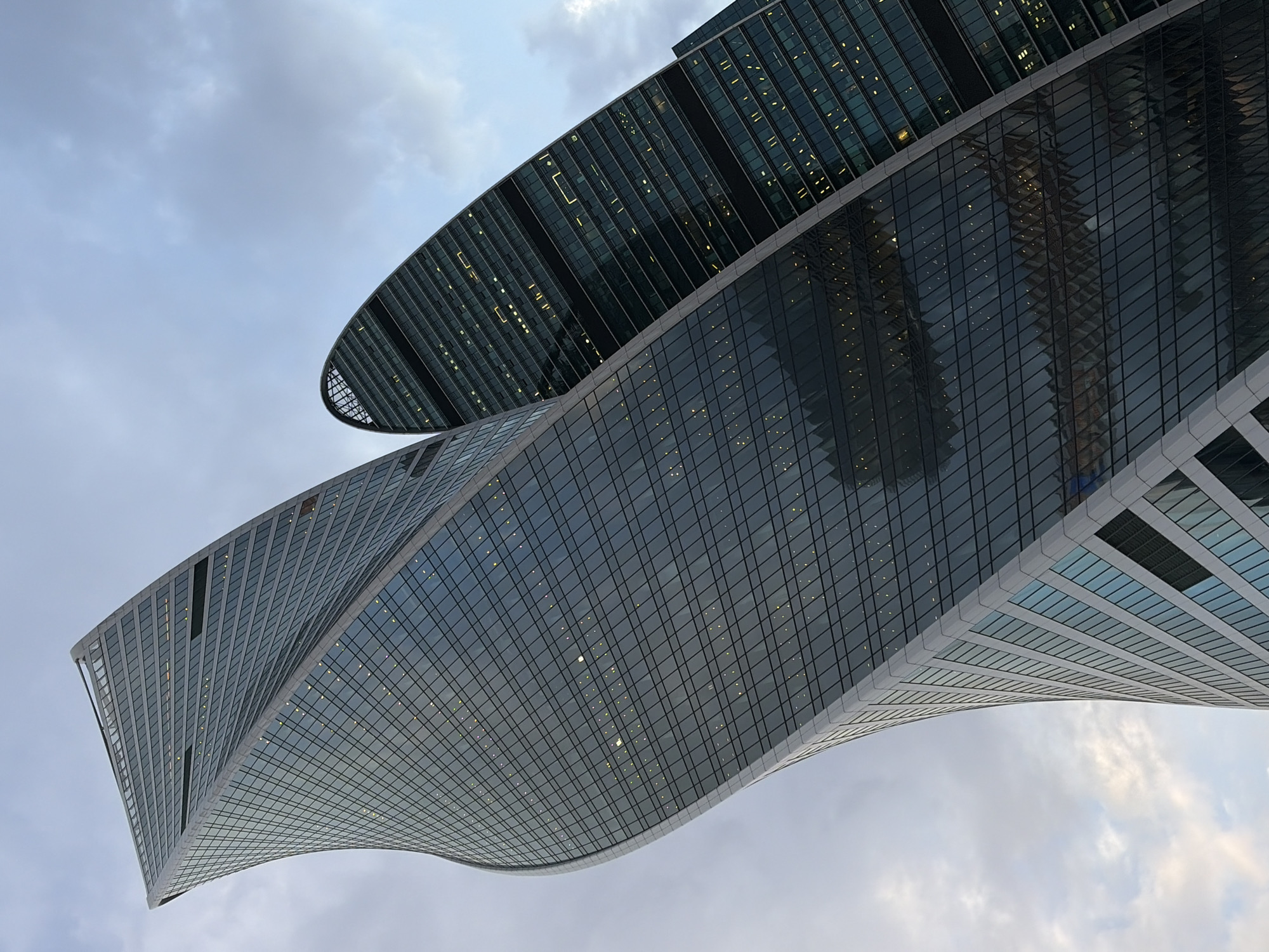The image shows two modern skyscrapers in Moscow, Russia, viewed from a low angle. The lower building has a twisting design, with reflective glass panels forming its exterior. The upper building has a flat, oval shape. The sky is overcast.