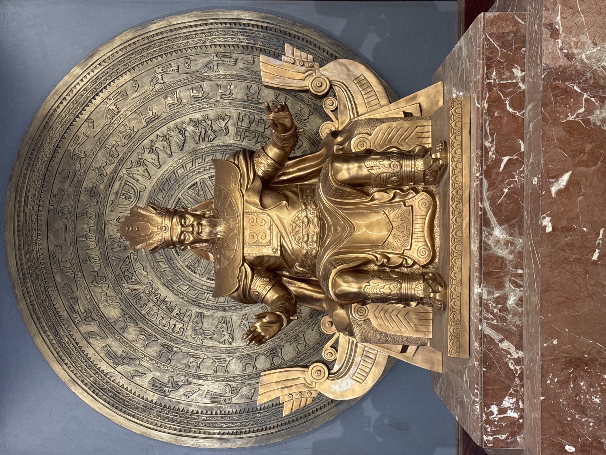 A full shot shows a golden statue of a king or emperor seated on a throne. The statue is positioned in front of a large, circular bronze disc decorated with intricate patterns and figures. The throne itself has a boat-like structure on either side. The statue is wearing a crown, ornate armor, and flowing robes. The king has a beard and is gesturing with his hands. The statue stands on a multi-tiered marble pedestal. The background is a plain blue wall. This statue is located at the Hung Kings' Temple complex in Hy Cương, Vietnam.
