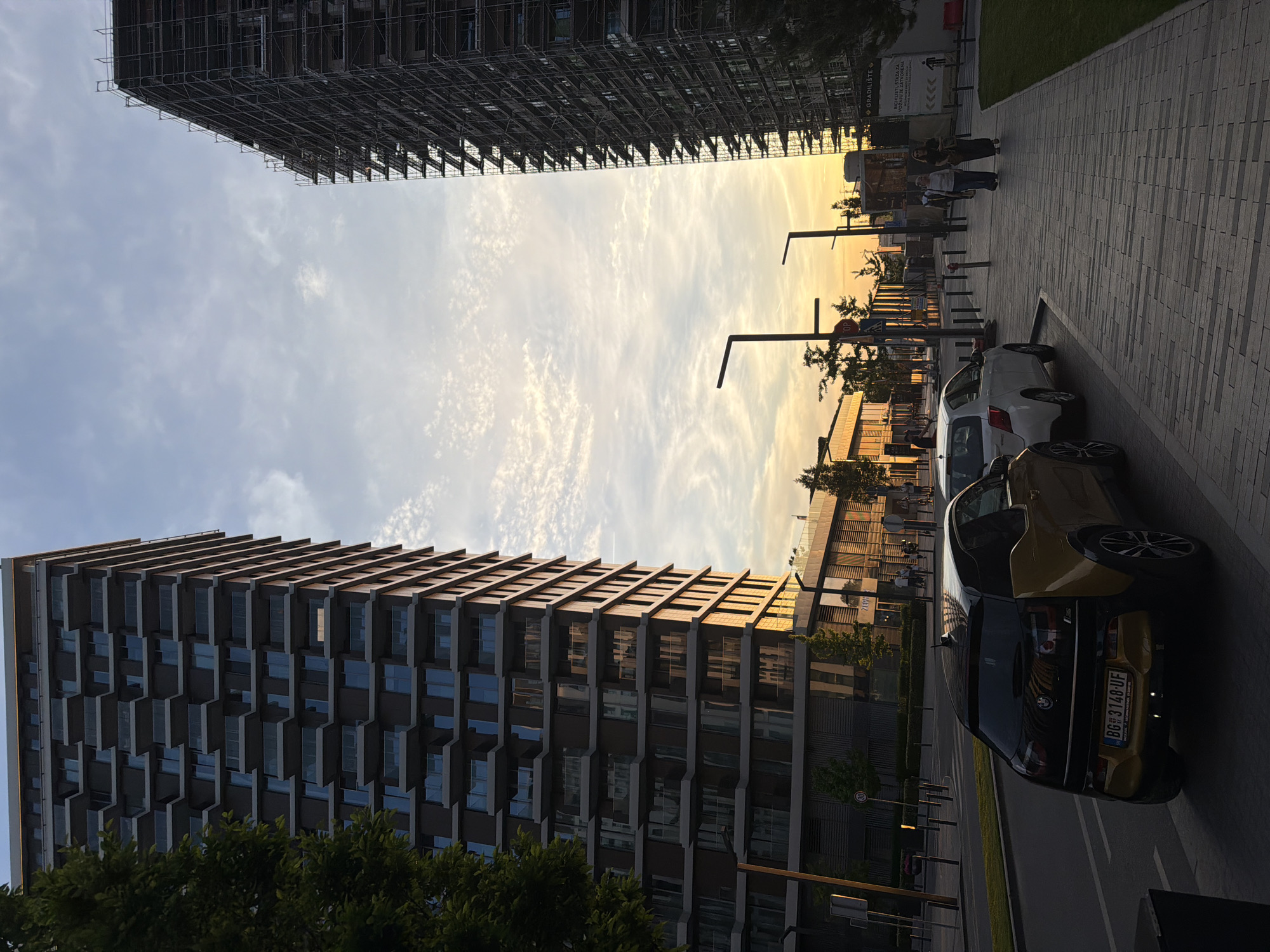 The image depicts a street scene in Belgrade, Serbia, featuring modern architecture, vehicles, and pedestrian activity. The left side of the frame showcases a tall, contemporary building with a repeating pattern of projecting balconies. A building under construction, enveloped in scaffolding, appears at the top of the image.
The sky overhead is filled with streaky clouds. Along the right side of the street are parked cars, including a dark-colored BMW, a silver car, and a yellow car.
Street lamps line the sidewalk where a small group of people walks in the distance. Other urban elements such as trees, street signs, and a paved walkway are visible. The scene is bathed in the golden light of either sunrise or sunset.