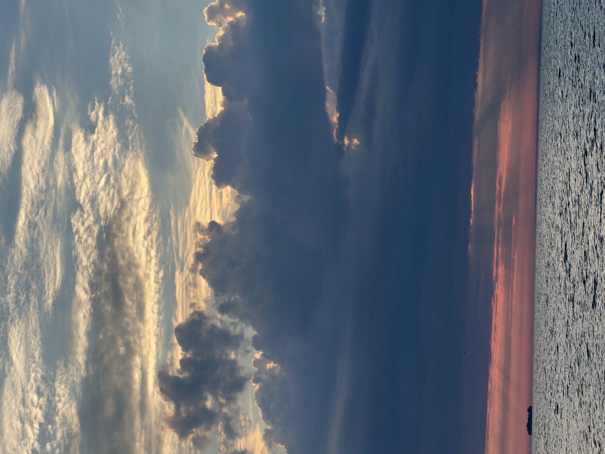 The image depicts a serene seascape at sunset, with a vast expanse of water meeting a dramatic sky. The water is a deep blue, rippled with gentle waves. Along the horizon, the sky transitions from a soft pink and orange near the water's edge, suggesting the last rays of the setting sun, to darker, brooding clouds above. These clouds are a mix of deep blues and grays, with some lighter, illuminated edges where the sun's light breaks through. A small, dark silhouette of what appears to be a ship or a barge is visible on the left side of the horizon, seemingly a lone traveler in the immense seascape. The overall impression is one of natural beauty and tranquility, characterized by the interplay of light and shadow in the sky and the calm surface of the water. The location specified, Borovoe, Russia, is a resort town known for its natural beauty, which aligns with the scene presented in the image.