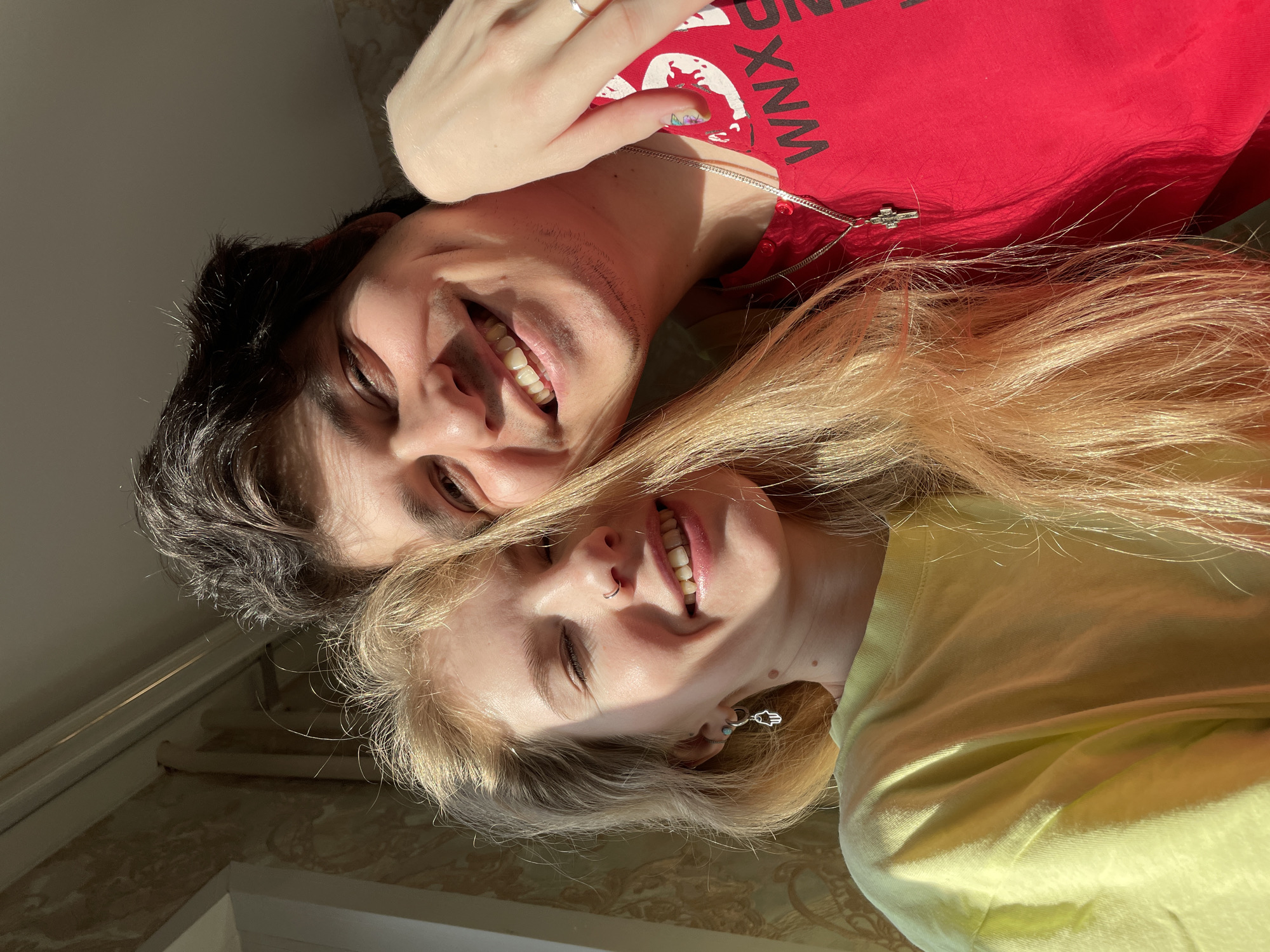 A selfie depicts two smiling individuals, a man and a woman. The man, positioned above, has dark hair, fair skin, and is wearing a red t-shirt with white lettering. A hand, possibly belonging to the woman, is visible near his neck, adorned with a ring on a finger and what appears to be nail art. He wears a silver chain around his neck with a cross pendant. The woman, positioned below, has blonde hair and a fair complexion. She wears a yellow t-shirt. She is wearing a hoop nose ring in her left nostril and an earring with a Hamsa pendant. The background suggests an indoor location, possibly in Izhevsk, Russia, with light-colored walls, decorative moldings and the edge of a window. No specific landmarks are discernible.