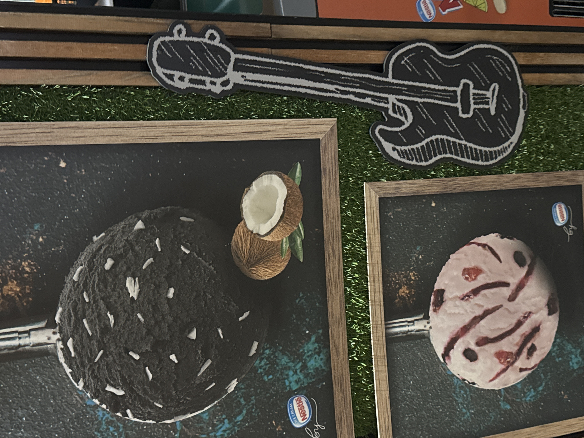 The image shows a display in an indoor setting, featuring two framed pictures of ice cream scoops. The scoop on the left is dark in color with white flecks and a coconut, while the scoop on the right is light-colored with dark streaks. Above the frames, there's a drawn cutout of a guitar. The guitar and frames rest on artificial grass, which is fixed to a set of wooden bars. The scene is likely related to an advertisement or promotion for ice cream, indicated by the "Nestle" logo on the pictures. The location in Veliko Tarnovo, Bulgaria, is not directly apparent from the image.