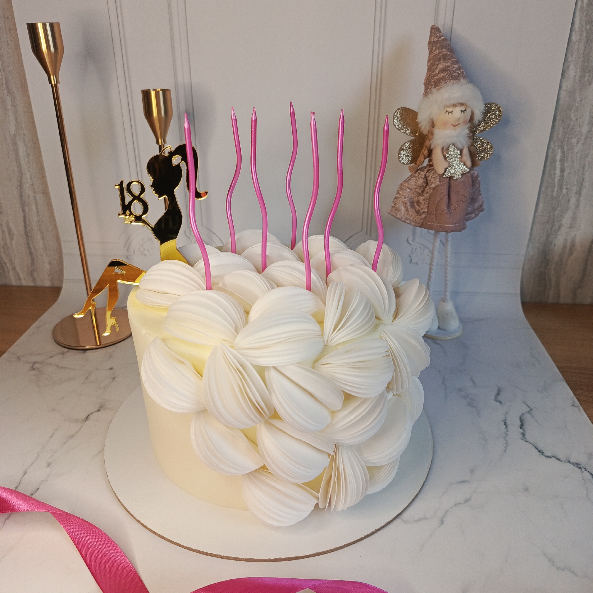 A birthday cake is centered on a white circular base. The cake is adorned with white petal-like frosting and eight pink candles. A gold "18" cake topper with a female silhouette in high heels is positioned on the left side of the cake. Beside it are two gold candle holders. To the right of the cake is a decorative doll with a pointed hat, wings, and a dress. A bright pink ribbon partially enters the frame at the bottom left. The background is a marble-patterned surface, which transitions to a wooden surface at the top left. The photo was likely taken indoors in Марковский, Russia.