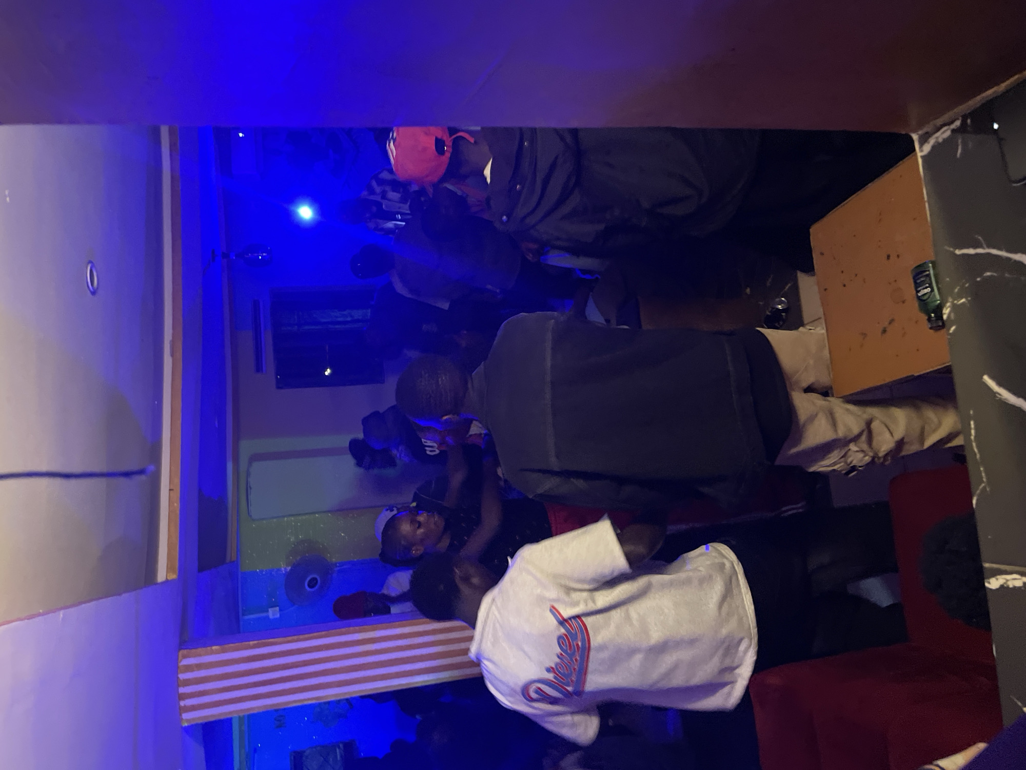 The image captures a dimly lit interior of what appears to be a social gathering or club in Durumi, Nigeria. The environment is illuminated by vibrant blue and purple lighting, with some areas casting strong shadows. A prominent pillar decorated with orange and white stripes stands to the left, contributing to the club-like ambiance. Several individuals are present, mostly in the mid-ground and background, engaged in various activities.
In the foreground, a person wearing a white t-shirt with a "Diesel" logo is facing away from the camera, their back to the viewer. To their right, another person is visible wearing a dark jacket. A table with a speckled surface is in the lower right, with a small green bottle on it.
In the mid-ground, a woman in a black top is dancing or interacting with others. Other individuals are seen in the background, some with their faces obscured by darkness or the angle of the shot. The overall scene suggests a lively atmosphere, typical of a night-time entertainment venue.