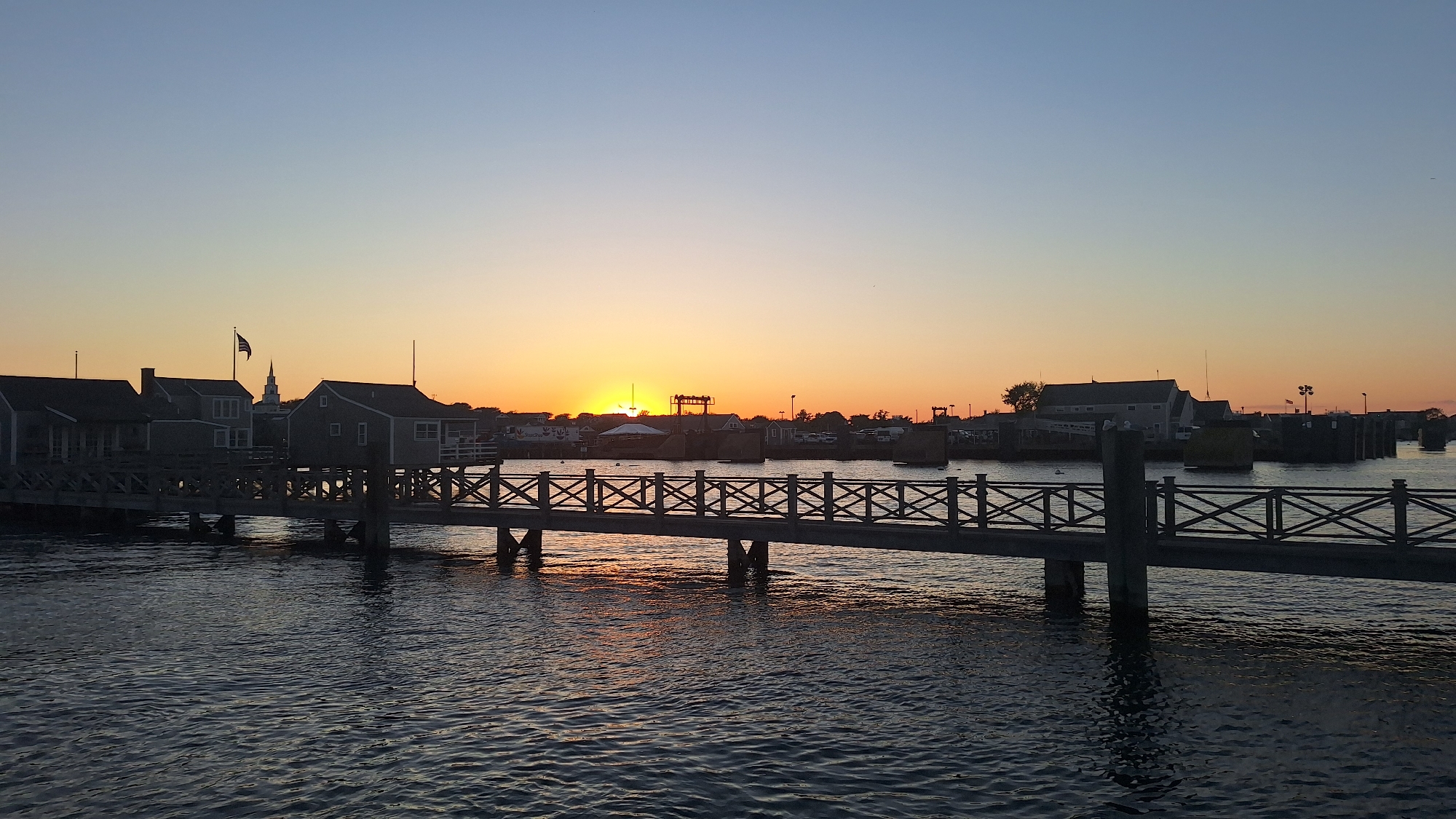 The image depicts a sunset scene over a harbor on Nantucket, United States of America. In the foreground, a wooden pier with a decorative railing stretches across the water. The pier is supported by pilings that are partially submerged in the dark, rippling water. In the middle ground, several buildings with grey exteriors and white trim are visible along the waterfront, some appearing to be boathouses or shops. An American flag flies from a pole in front of one of the buildings. Beyond the buildings, the sun is setting on the horizon, casting a warm orange and yellow glow across the sky. Silhouettes of trees and structures are visible against the vibrant sky. In the distance, more buildings and piers are faintly discernible. The overall impression is a peaceful harbor scene at dusk.