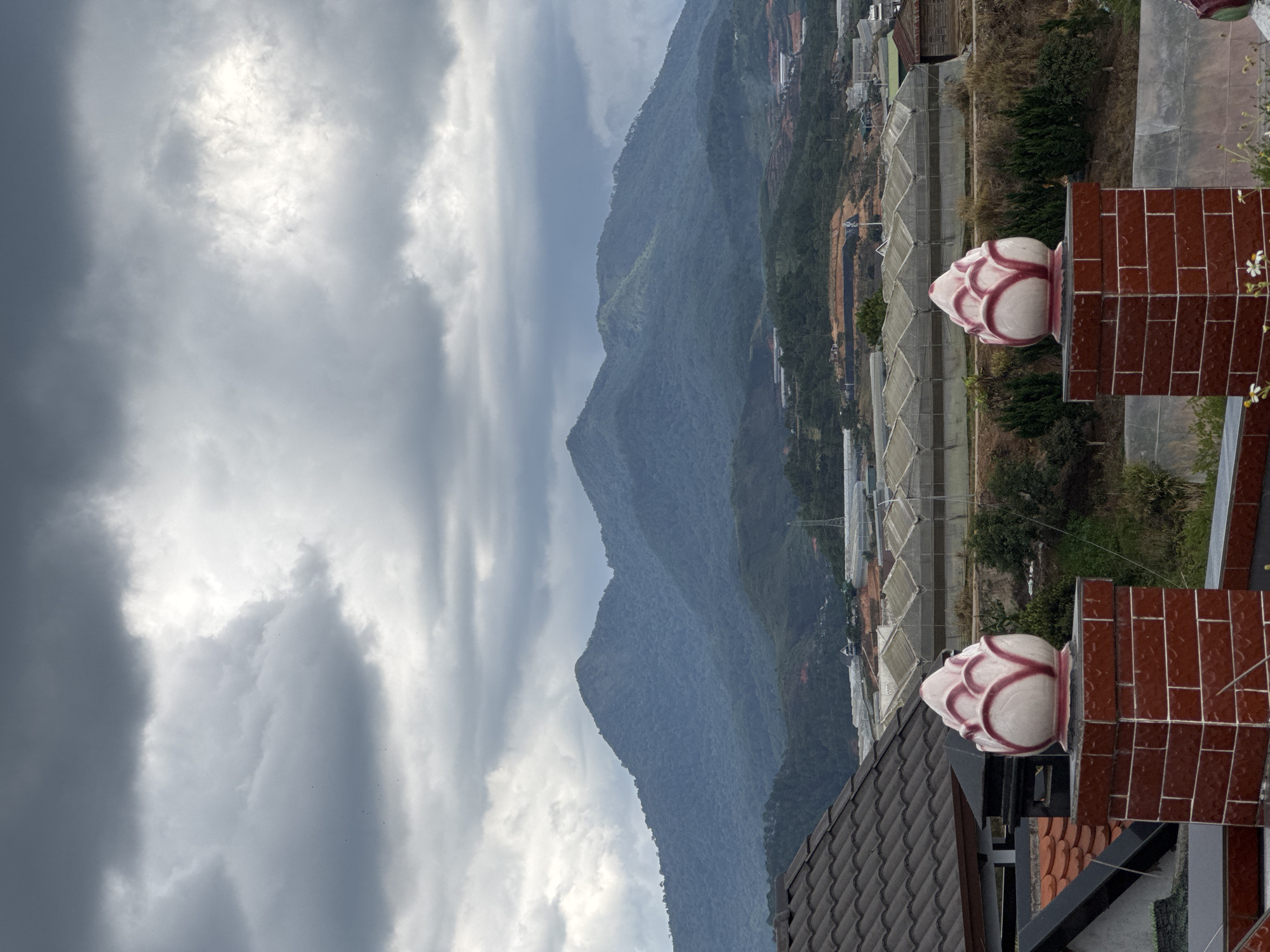 The image is taken from an elevated perspective, likely a rooftop, overlooking a mountainous landscape in Lạc Dương, Vietnam. The sky is overcast with heavy, grey clouds. In the mid-ground, forested mountains stretch across the horizon. Below the mountains, a valley contains numerous greenhouses with translucent covers, suggesting agricultural cultivation. In the foreground, architectural elements are visible: red-tiled structures and rooflines, with decorative, flower bud-shaped ornaments.