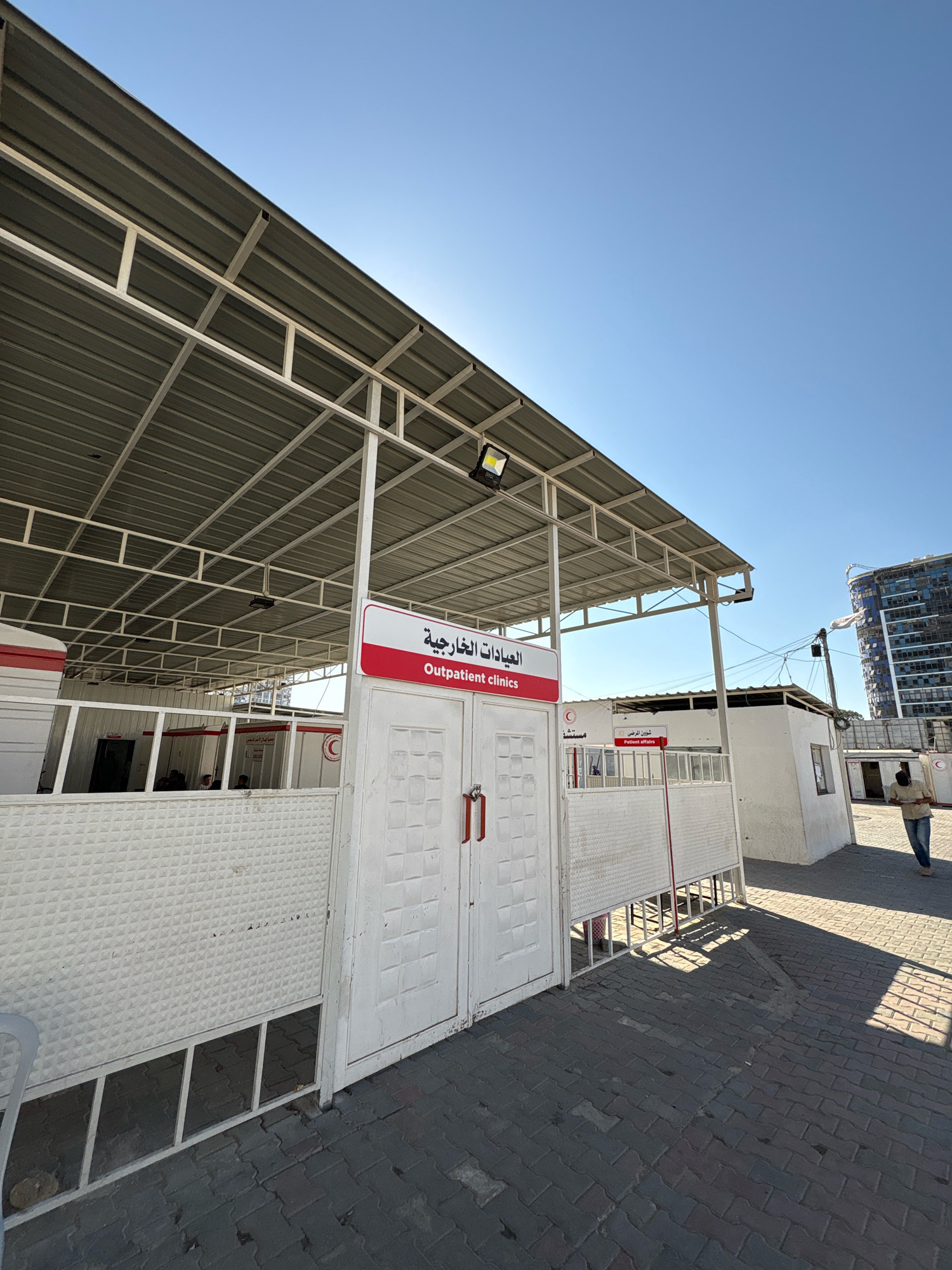 This is an exterior, eye-level, medium shot of an outdoor clinic facility, likely in Gaza City, Palestinian Territories. The primary subject is a white clinic building with a red and white sign that reads "Outpatient clinics" in English and Arabic. The building has white double doors with red handles. To the left of the doors is a white corrugated metal fence. To the right of the doors is a white, boxy structure with a smaller red and white sign that reads "Patient affairs" in English and Arabic, above an emblem of the crescent moon and red cross. A person wearing a light-colored shirt and dark pants walks away from the camera on the paved ground to the right. In the background, a tall, modern building with blue glass windows is visible against a clear blue sky. The clinic appears to be set up with a metal roof structure supported by white metal poles, covering an open-air waiting area. The ground is paved with dark grey interlocking pavers. The lighting suggests it is daytime with bright sunlight.