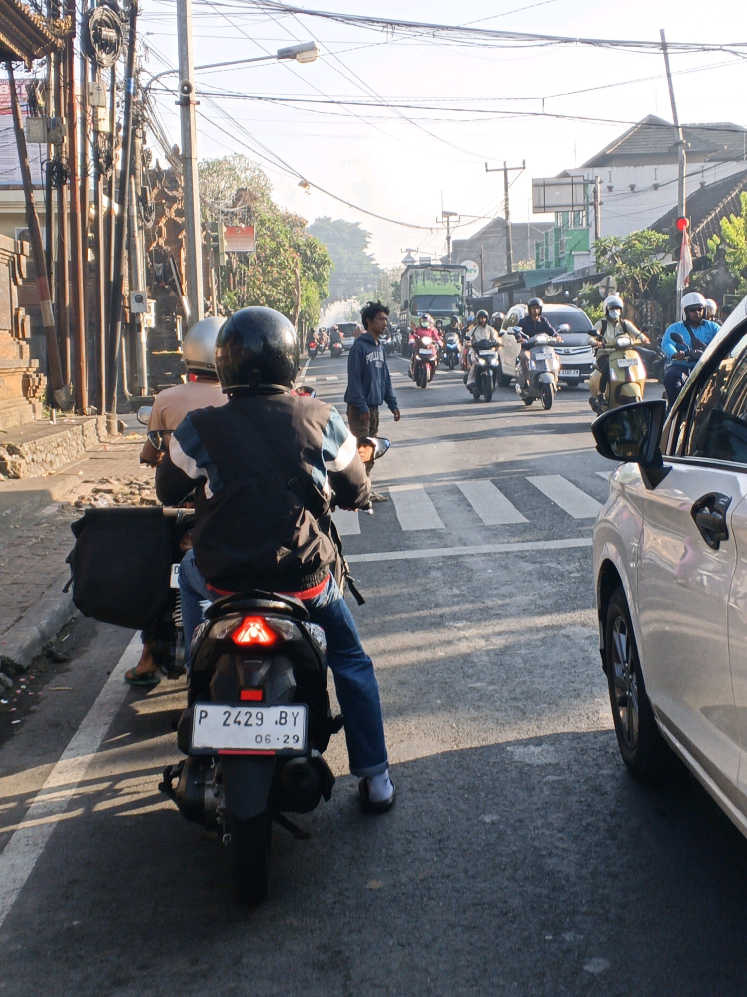 The image captures a street scene in Abiansemal, Indonesia, depicting a traffic jam with numerous motorcycles and a few cars. The time of day appears to be late afternoon, judging by the long shadows cast across the asphalt road. In the foreground, a person wearing a black jacket and blue jeans rides a scooter, their back to the camera. The scooter's license plate reads "P 2429 .BY 06-29". To the left of the scooter, another scooter with a silver helmet is visible.
The road is paved and marked with a pedestrian crossing in the middle distance. The surrounding environment includes buildings with traditional Indonesian architecture, utility poles laden with electrical wires, and lush green trees. Several other individuals are seen on motorcycles, some wearing helmets, navigating the congested street. A white car is positioned on the right side of the frame, partially obscuring the view of the traffic ahead. The overall atmosphere is one of everyday urban life and transportation in Indonesia.