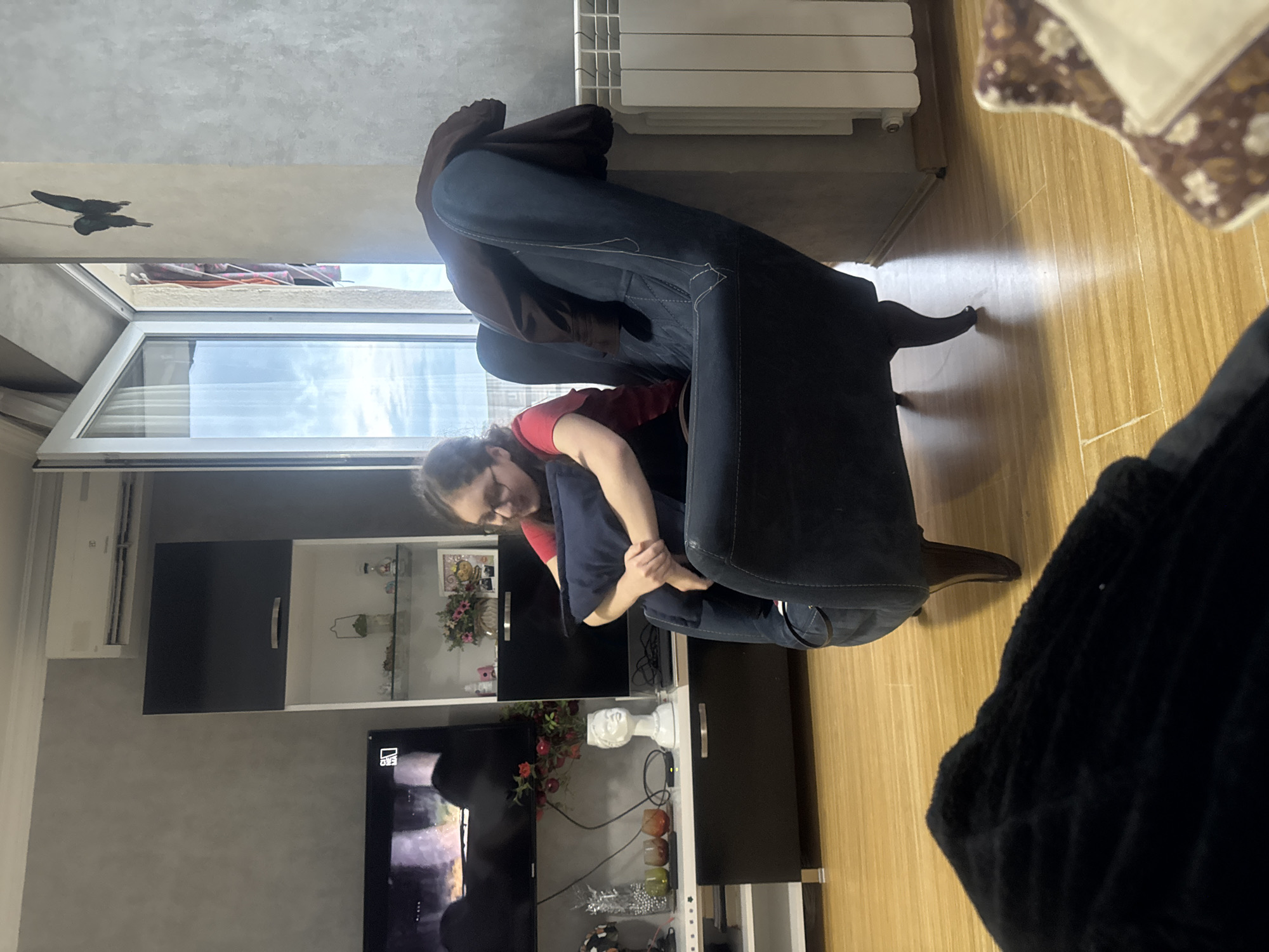 A woman wearing glasses and a red shirt is sitting on a dark blue armchair in a living room in Tbilisi, Georgia. The room has a large window with a view of the sky, a radiator, a television, and a display cabinet filled with decorative items. A dark blanket is visible in the foreground.