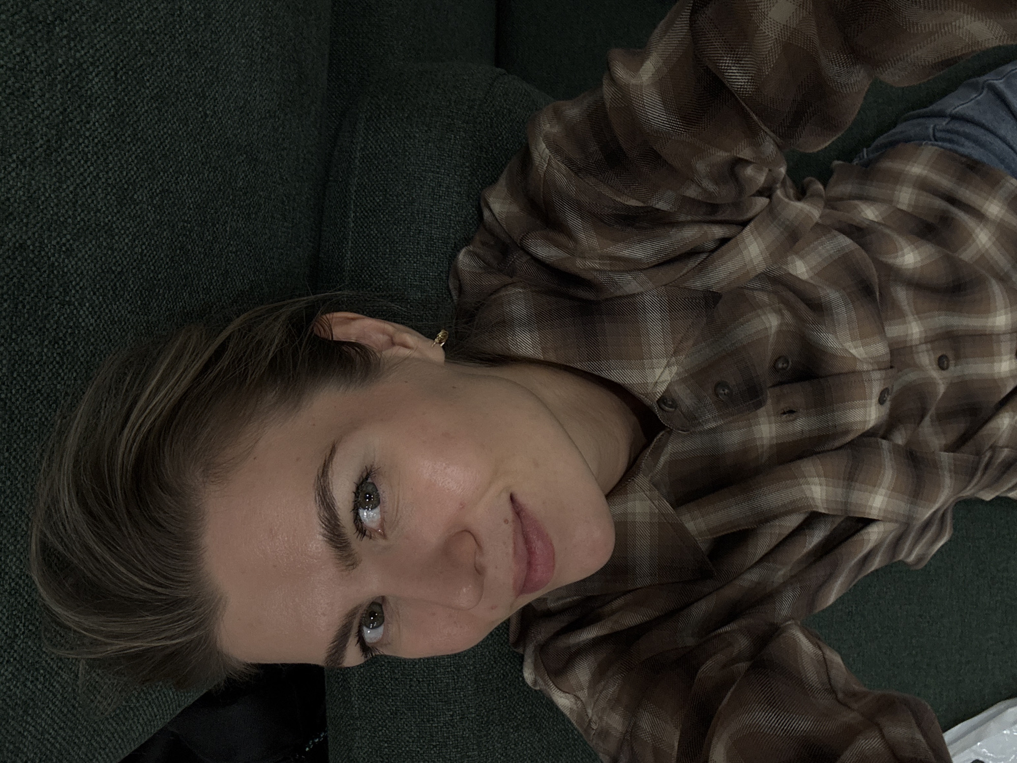 A woman is lying on a green couch, wearing a brown plaid shirt and blue jeans. She is looking up at the camera. The location is Unknown, Unknown.
