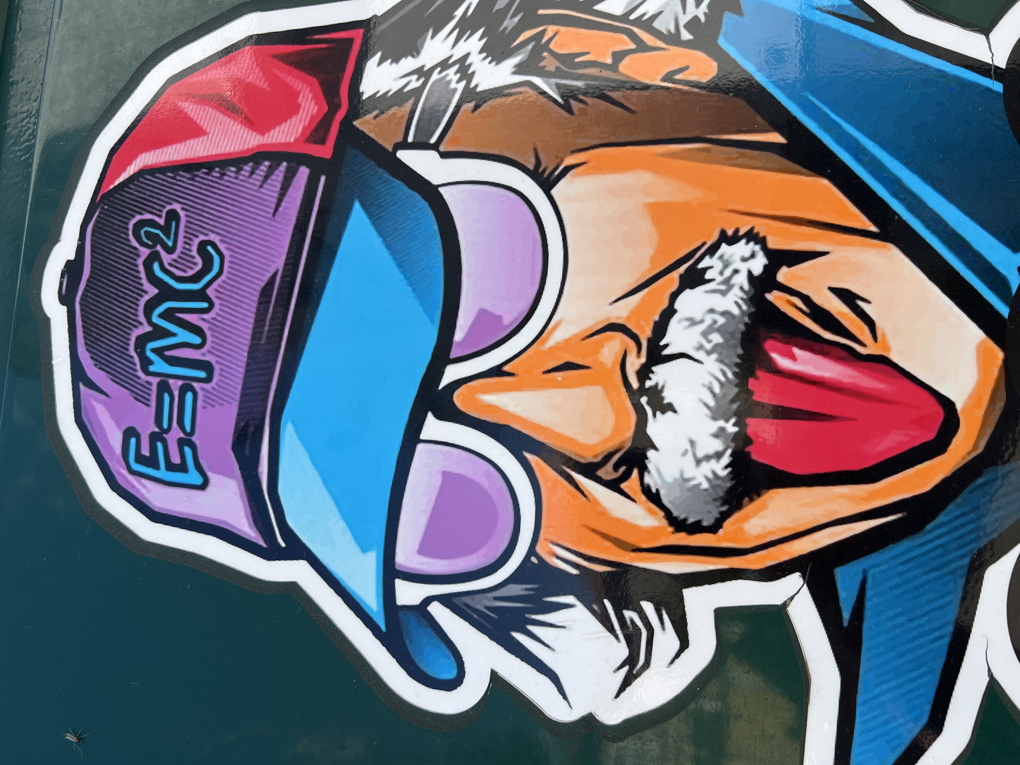 The image shows a sticker or painting depicting a caricature of Albert Einstein wearing a backwards baseball cap with "E=mc^2" printed on the side. He is wearing sunglasses with purple lenses and his tongue is sticking out. The background is dark green. The location is Burgas, Bulgaria (This information is based on source images from Burgas, Bulgaria).