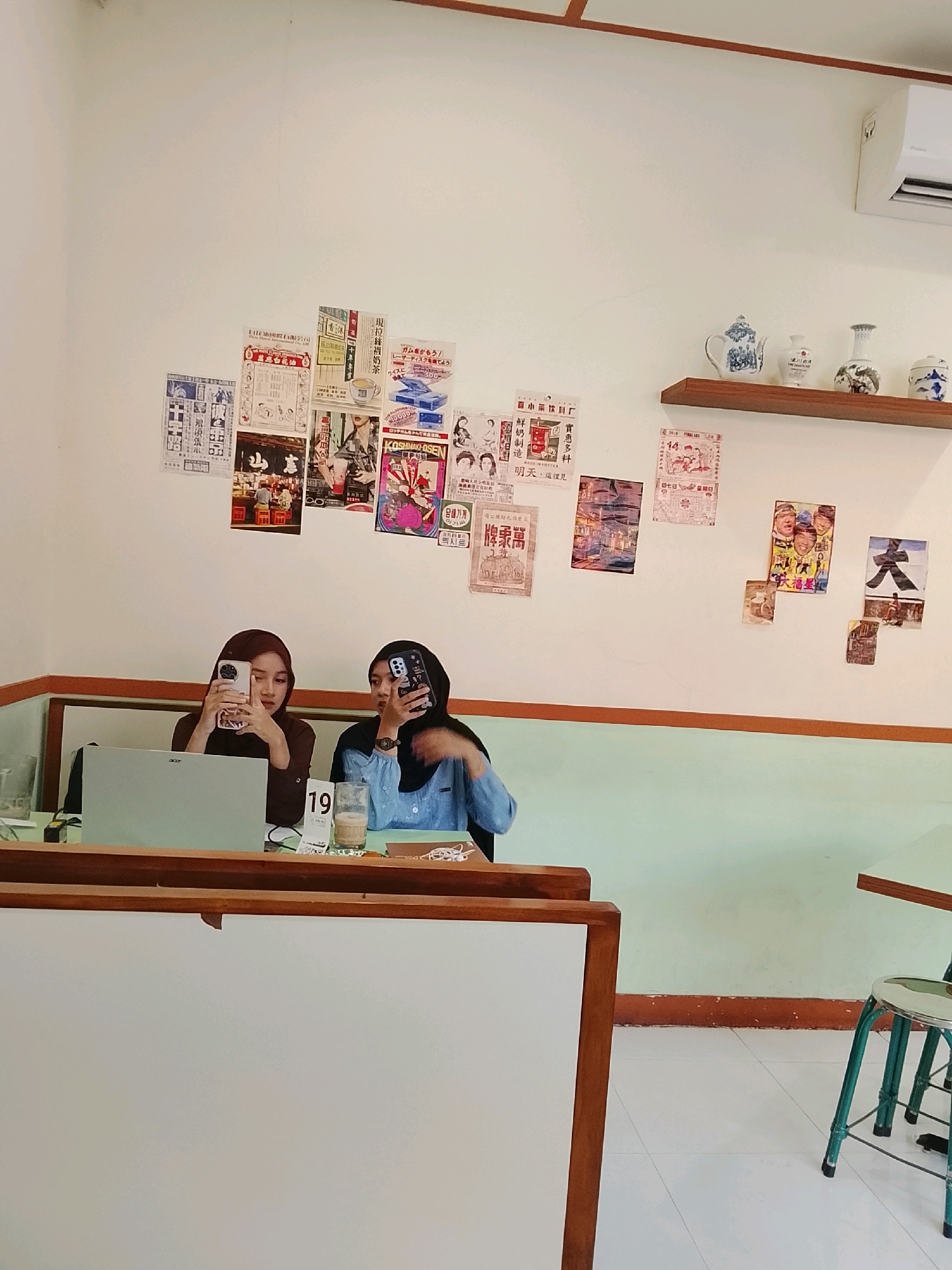 This is a photograph of two young women, both wearing hijabs, sitting at a table in what appears to be a cafe. The woman on the left is wearing a dark brown shirt and is looking at her phone, which she is holding up. She has a laptop in front of her. The woman on the right is wearing a light blue shirt and is also looking at her phone, which she is holding up. In front of her is a glass of iced coffee. A calendar showing the number 19 is visible between them. The wall behind them is decorated with various posters and pictures, some of which appear to be vintage. There are also two decorative ceramic vases and a teapot on a shelf above them. The cafe has light green walls and light-colored tiled floors. The overall impression is that of a casual, indoor setting. The image does not contain any specific visual elements that definitively place it in Surabaya, Indonesia.