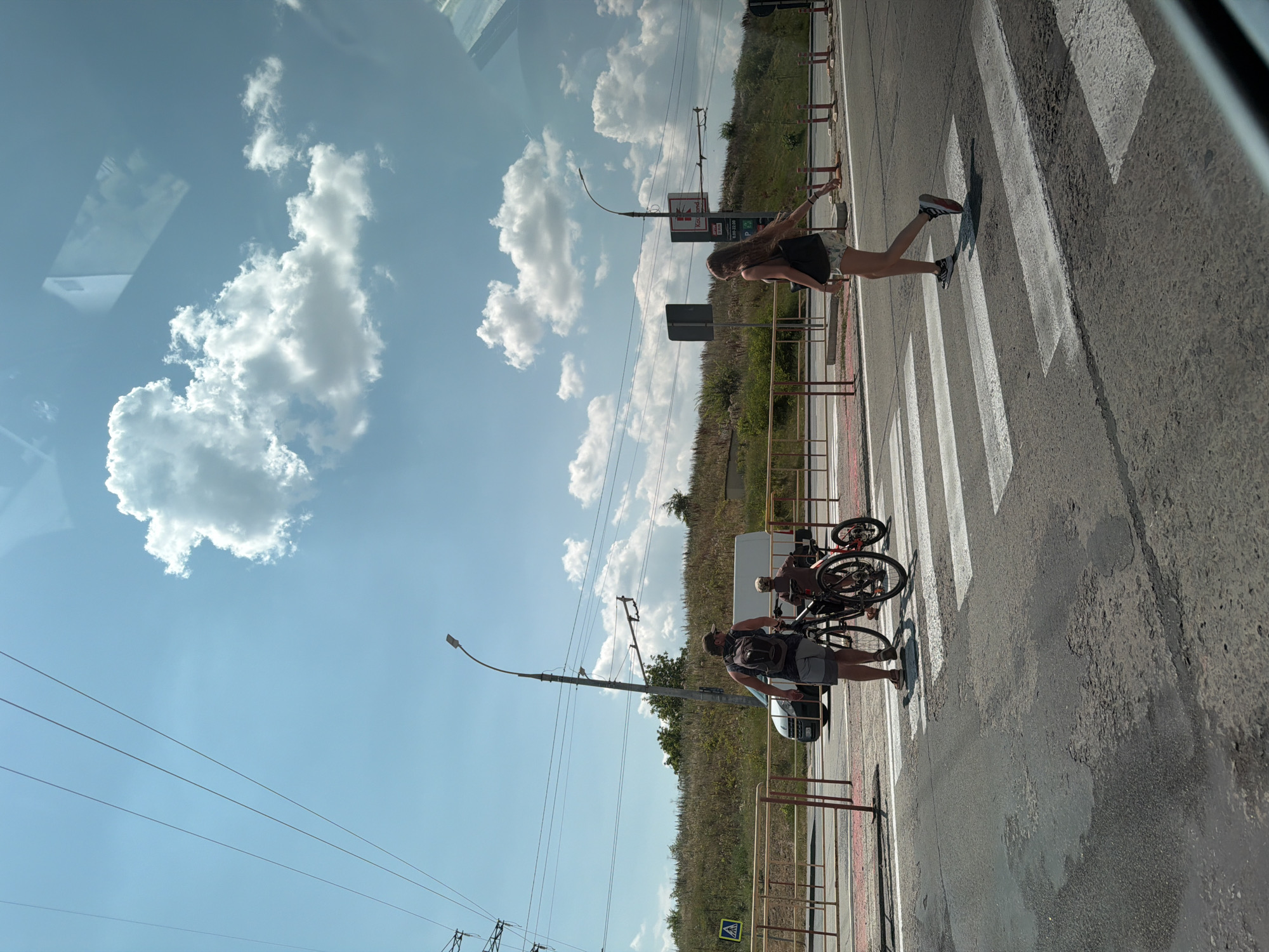 A bright, sunny day scene unfolds at a pedestrian crossing in Codru, Moldova. Three people are present. In the foreground on the left, a man with a backpack is holding a bicycle. Next to him, a child is seated on a smaller bicycle. To the right, a woman with long hair is walking across the pedestrian crossing. The background features a road with a van and a grassy embankment dotted with trees. Overhead, power lines and a street lamp are visible against a clear blue sky with scattered white clouds.