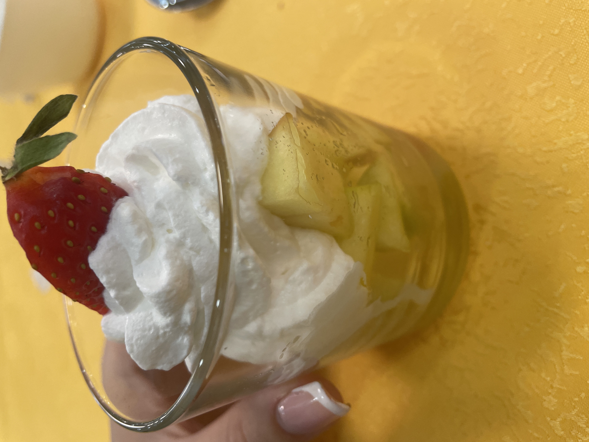 A close-up shot depicts a glass of fruit dessert held in someone's hand. The glass contains cubed fruit, possibly apples or melon, and is topped with a large mound of whipped cream. A single strawberry with green leaves is placed on the side of the glass, nestled against the whipped cream. The dessert rests against a yellow textured surface. The photo was taken indoors in Starozagorski bani, Bulgaria. The hand holding the glass has a manicured fingernail with a white tip.