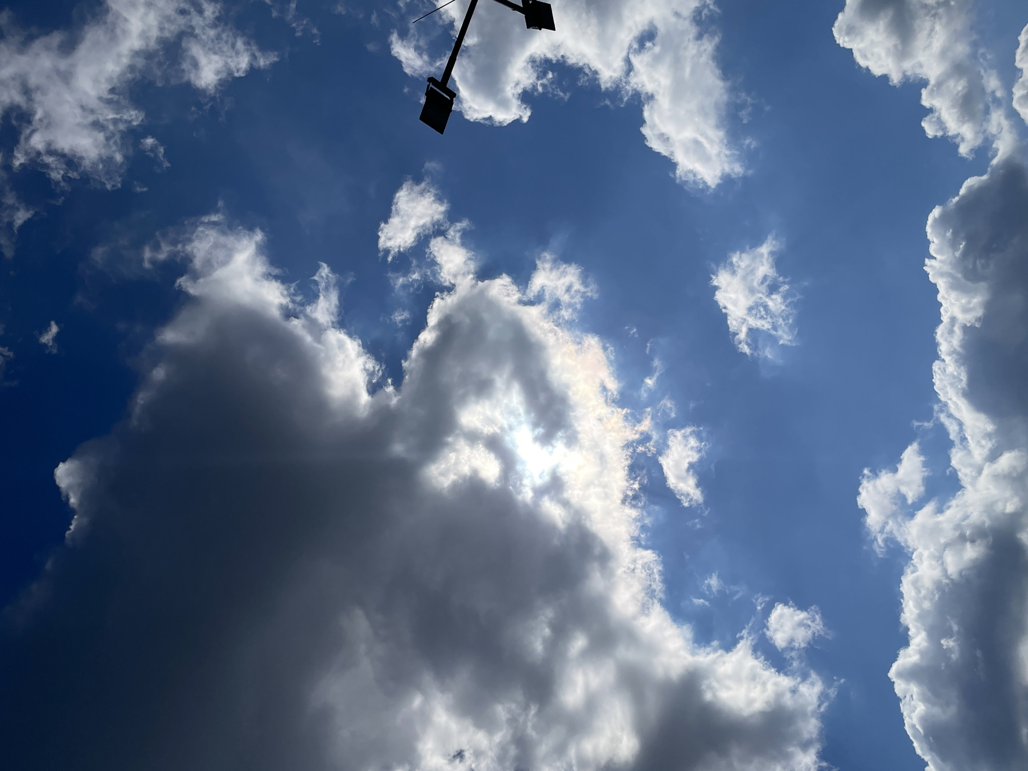 This is a vertical, low-angle shot of the sky, with a large dark cloud dominating the left side of the frame, partially obscuring the sun. Smaller, white cumulus clouds are scattered across the bright blue sky. On the right side of the image, a silhouette of a streetlamp with two lamps is visible against the sky. The scene is outdoors, with no specific indication of a city or location.