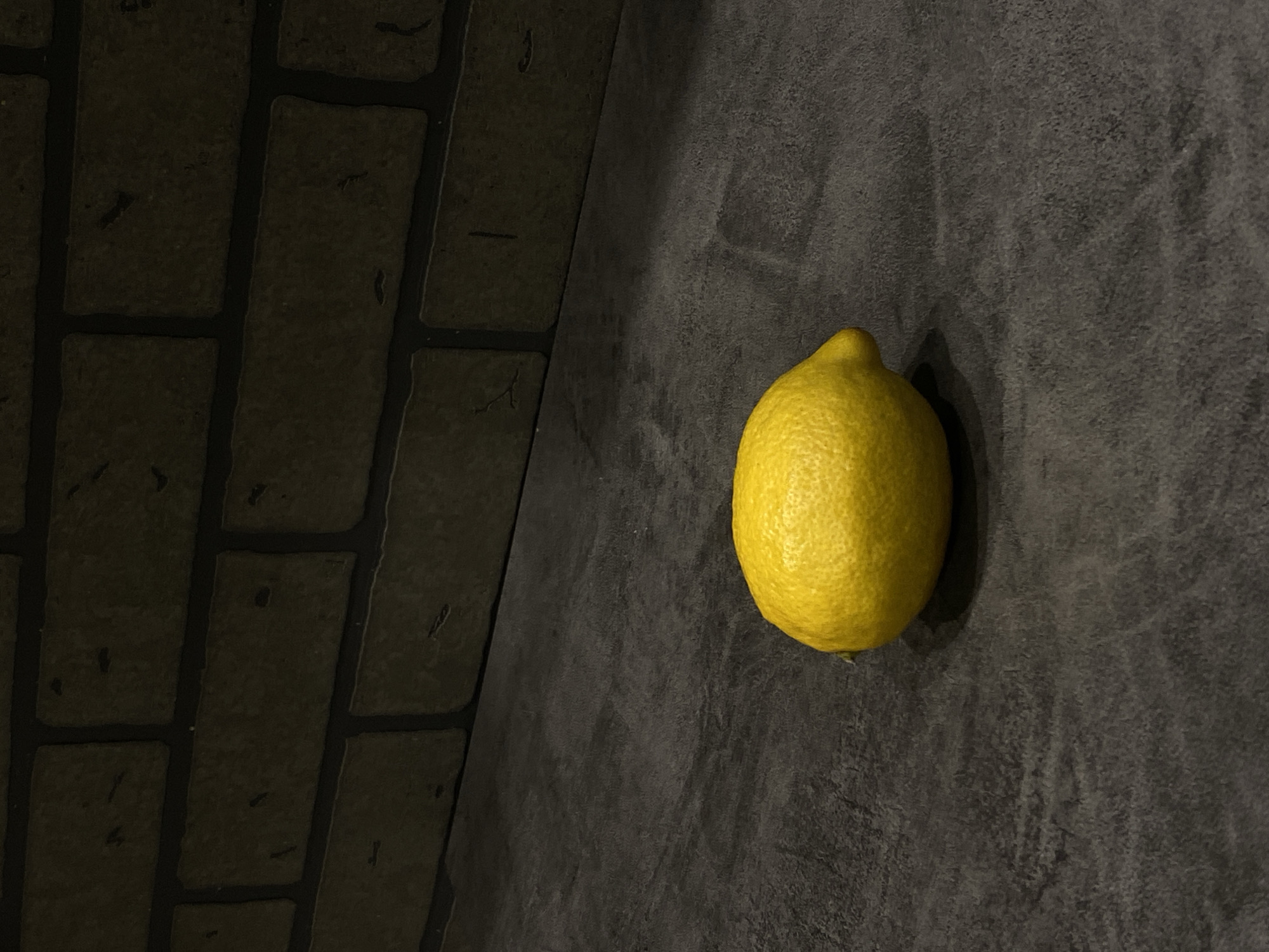 A single lemon rests on a dark gray surface that transitions to a brick-like pattern in the upper left quadrant. The lemon's yellow color contrasts sharply with the somber tones of the background. The scene is sparsely lit, with the lemon capturing the majority of available light. There are no discernible people, interactions, or clear environmental cues indicating the specific location of Sochi, Russia.