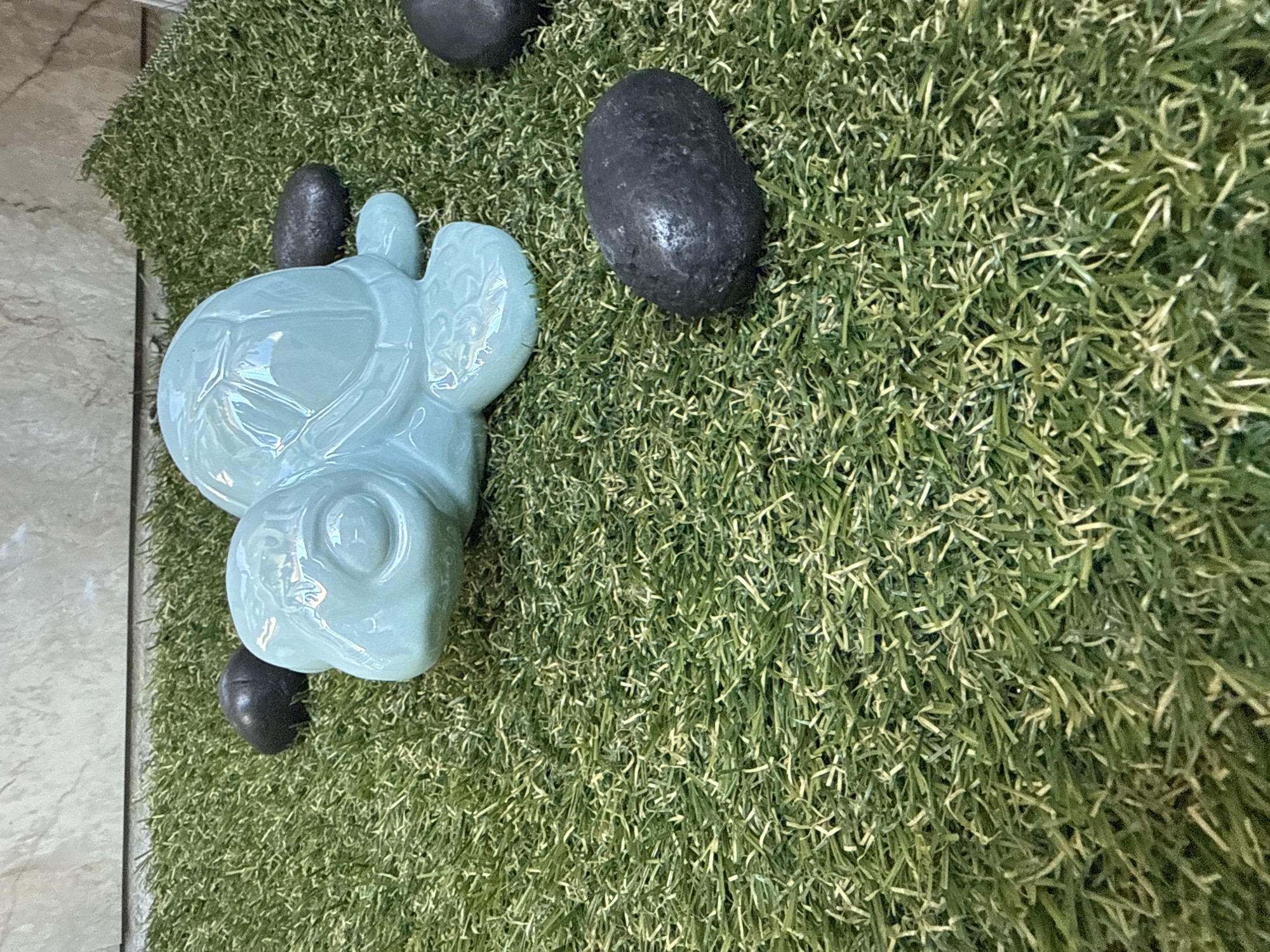 A light blue ceramic turtle rests on a patch of artificial grass. Several dark, smooth stones are scattered around the turtle. The background is a textured wall made of light-colored rectangular tiles. The scene is indoors, possibly in a decorative setting within a home or business. The image does not provide any information about the city of Sofia, Bulgaria.