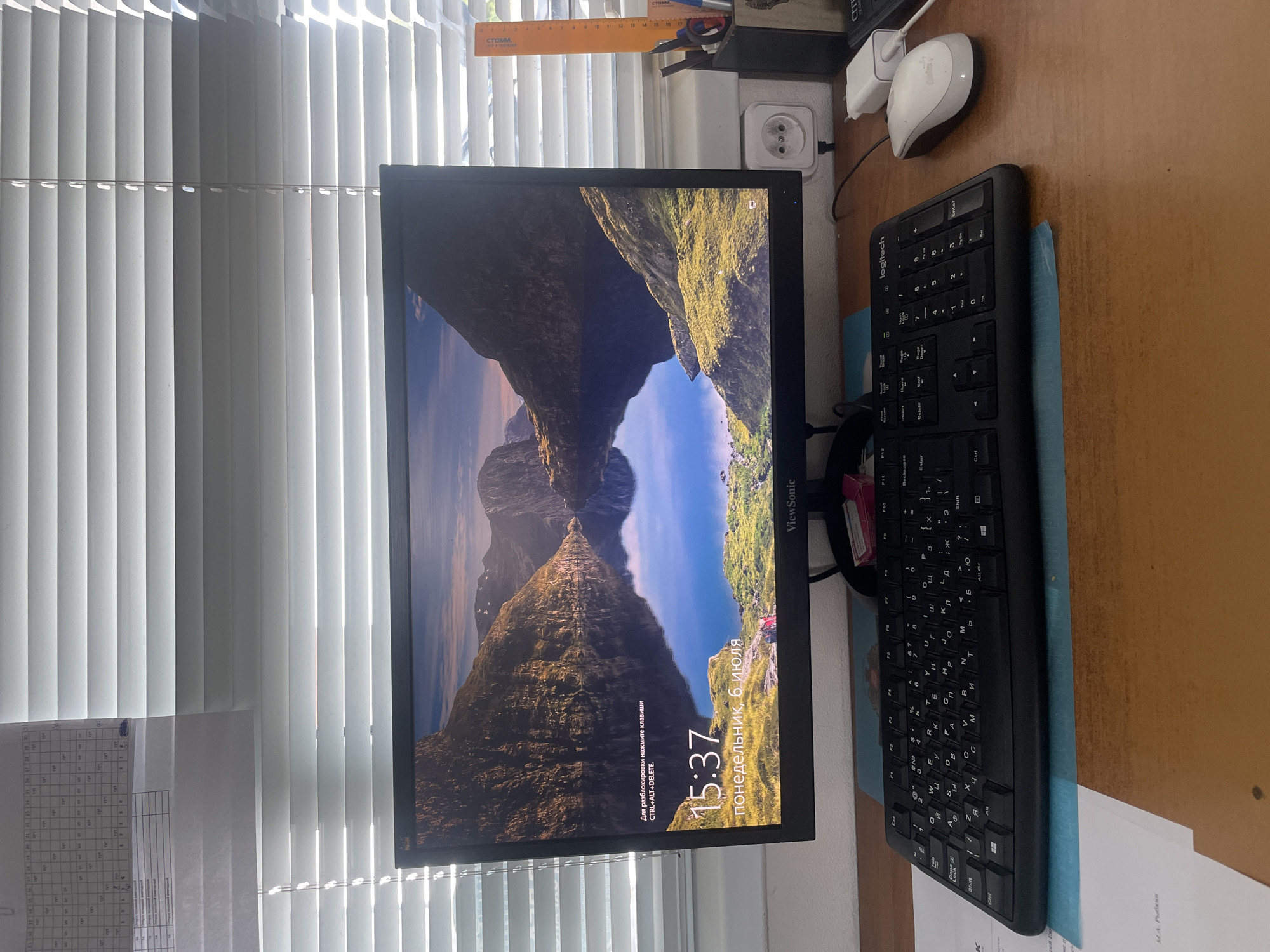 The image shows a computer setup on a wooden desk. A large monitor displays a scenic landscape of mountains and a lake, with the time "15:37" and the date "понедельник, 6 июля" (Monday, July 6) visible. Below the monitor is a black keyboard with both Cyrillic and Latin characters. A white computer mouse is to the right of the keyboard, and a white power adapter is plugged into an electrical outlet on the wall behind the monitor. The scene is illuminated by sunlight coming through horizontal blinds on a window. To the left of the monitor, a printed schedule or calendar is partially visible, attached to the blinds. To the right of the monitor, a white pencil holder contains pens, scissors, and an orange ruler. The image does not contain enough information to determine the location in the city of Salsk, Russia.